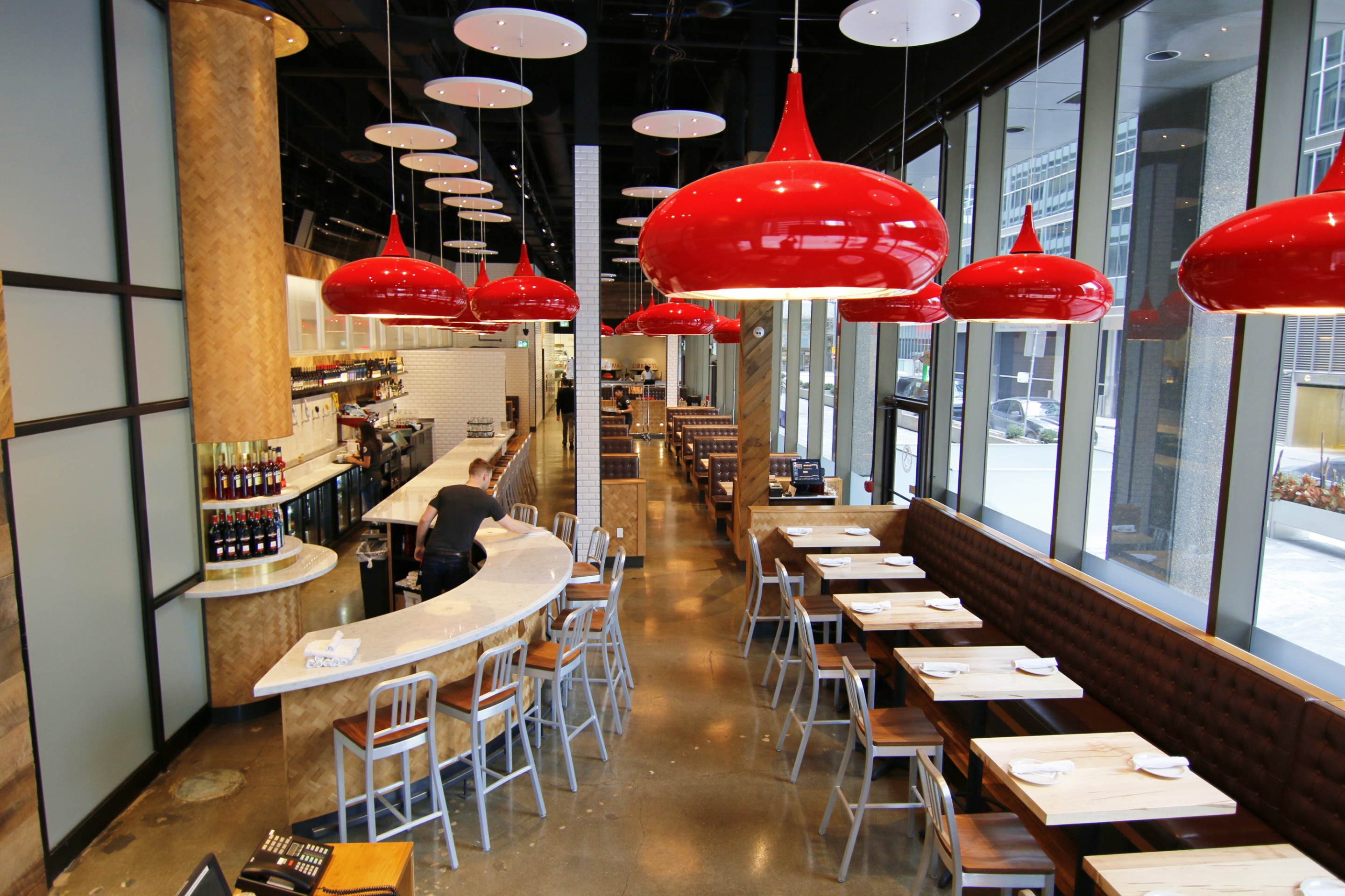 A modern restaurant interior with a curved bar, wood tables, and bench seating along large floor‑to‑ceiling windows. Red pendant lights hang from a dark ceiling, adding contrast to the bright, contemporary dining space.