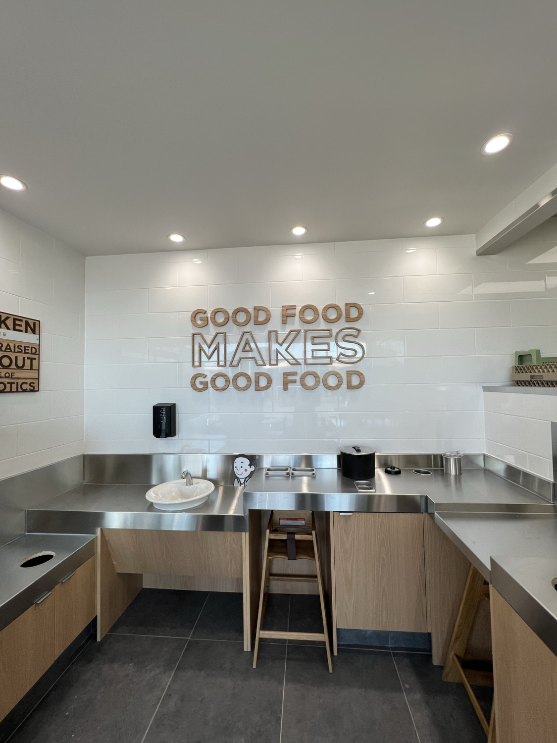 A clean, modern kitchen area with stainless steel counters, a sink, and light wood cabinetry. White tiled walls display the sign “GOOD FOOD MAKES GOOD FOOD” above the workspace, illuminated by ceiling lights.