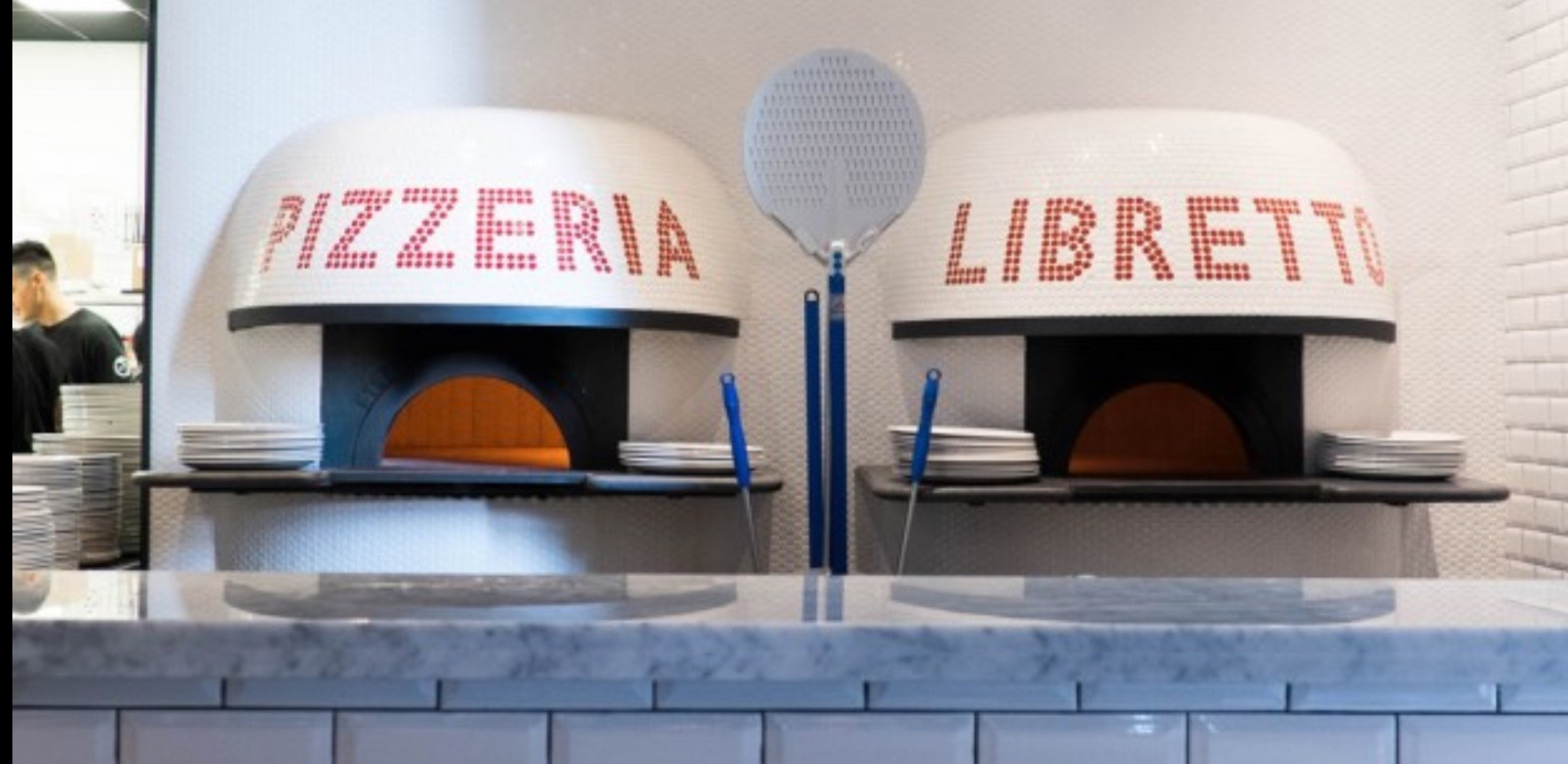Two white, dome-shaped pizza ovens labeled “Pizzeria” and “Libretto” sit behind a tiled counter. Stacks of plates and long-handled pizza tools are positioned between and beside the open oven mouths.