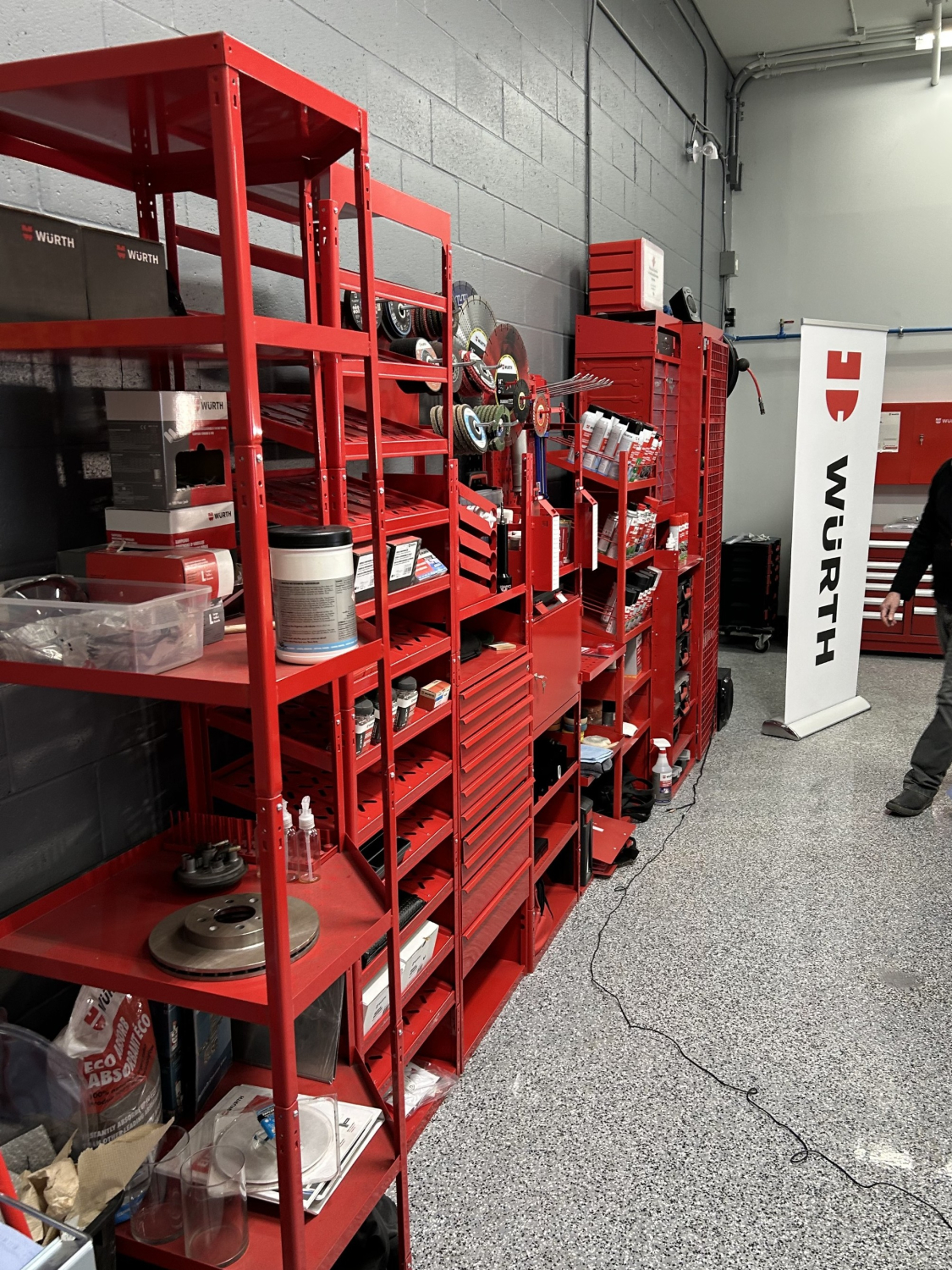 An interior view of a Würth supply area with tall red metal shelving filled with tools, parts, and containers. A white Würth banner stands beside the shelving in a clean, gray‑walled industrial room.