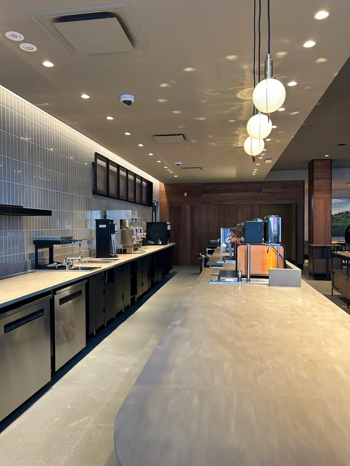 A modern café service counter with espresso machines, menu boards, and pendant globe lights overhead. Wood finishes, tiled walls, and stainless steel equipment create a clean, contemporary coffee bar interior.