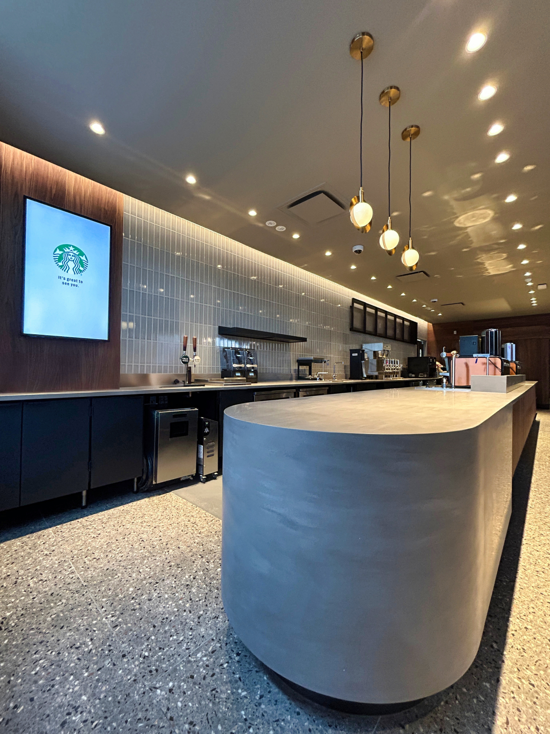 A modern café counter with a curved central work surface, espresso machines, and pendant globe lights overhead. Wood paneling, tiled walls, and recessed lighting create a clean, contemporary coffee bar interior.
