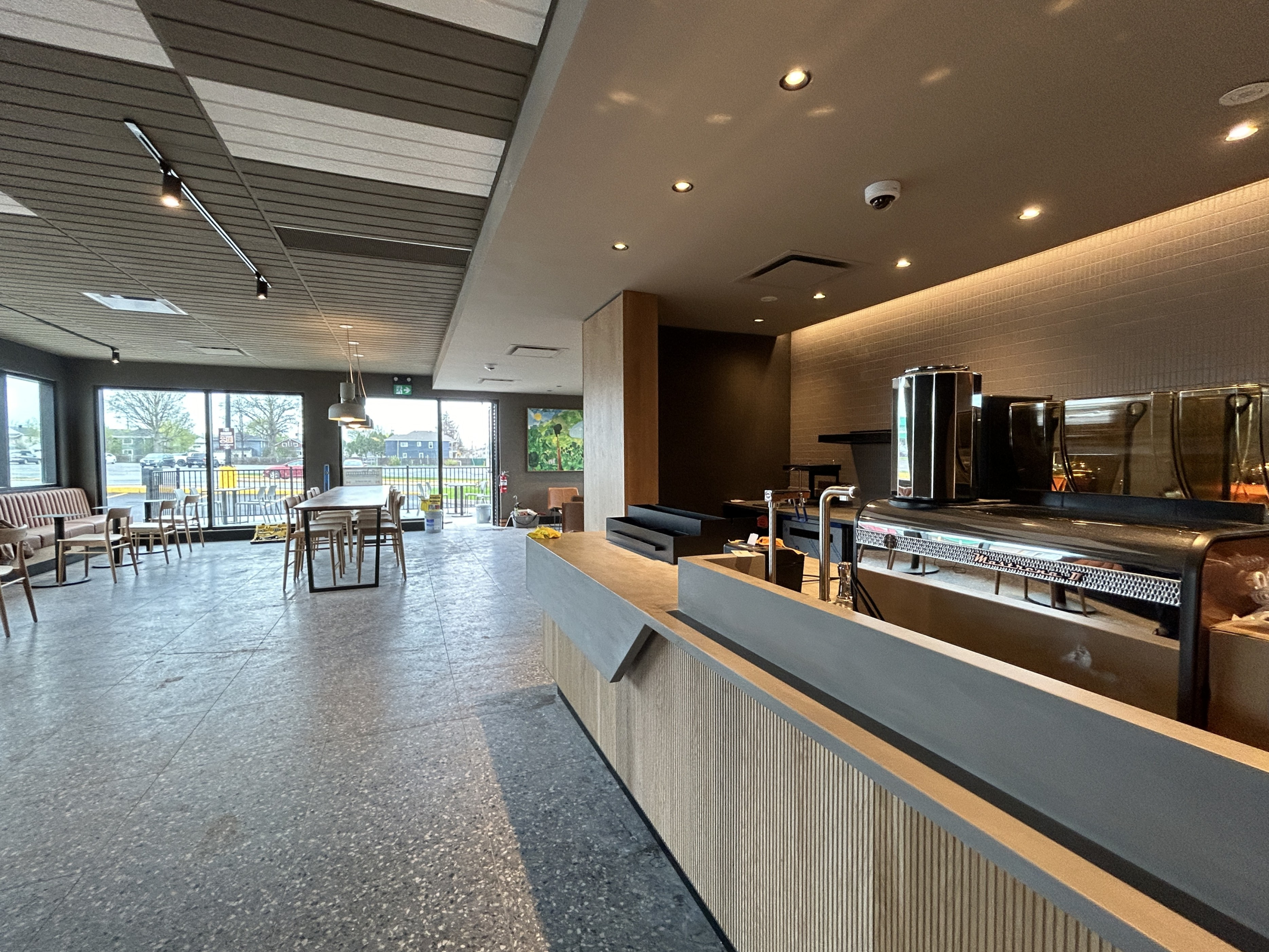 A modern café interior showing a coffee preparation counter with espresso equipment in the foreground and seating near large windows in the background. Polished floors, wood accents, and ceiling lighting create a clean, contemporary space.
