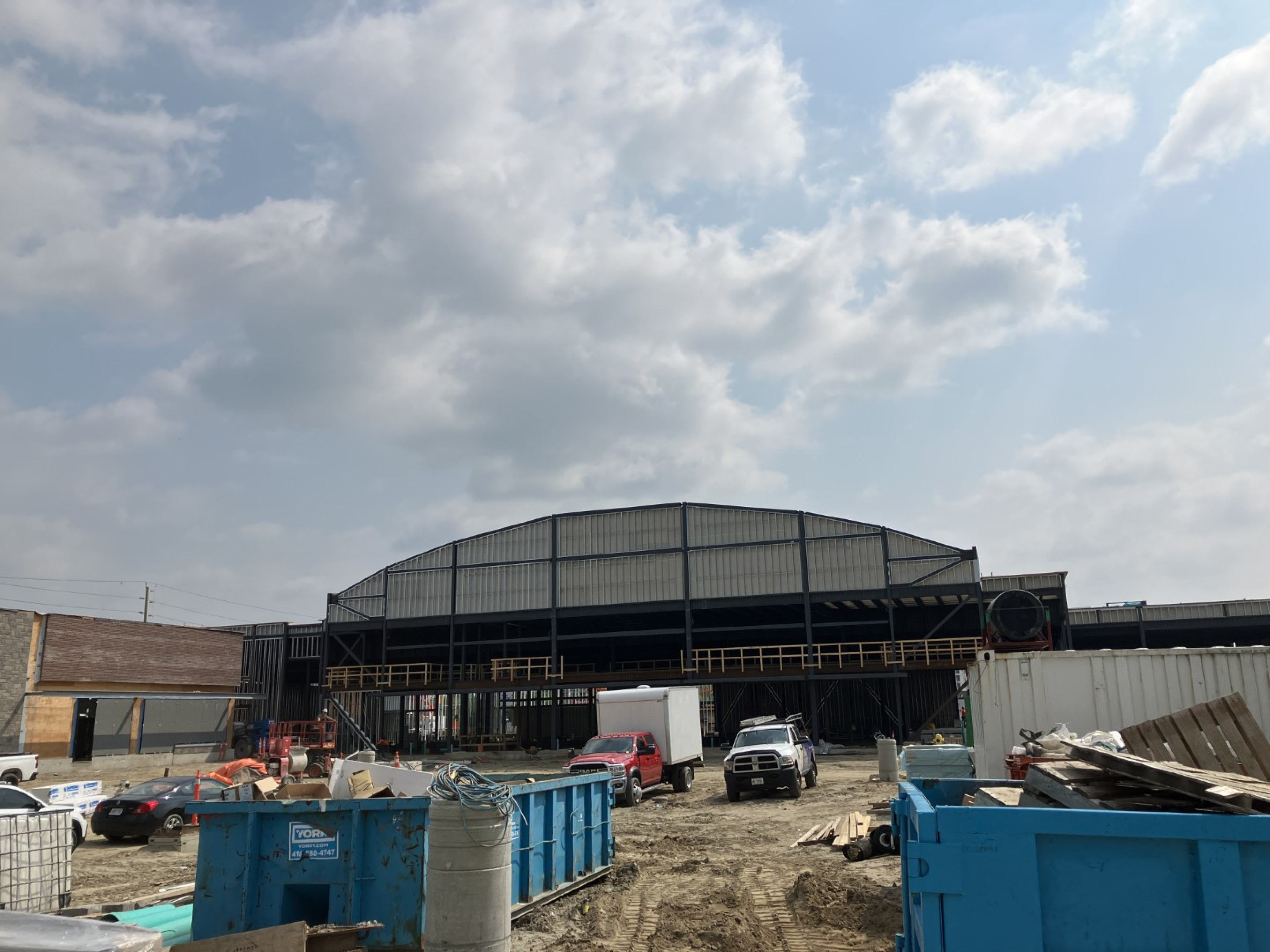 A large industrial building under construction with a steel frame and open facade sits on a dirt work site. Dumpsters, construction materials, trucks, and equipment are scattered in the foreground beneath a partly cloudy sky.