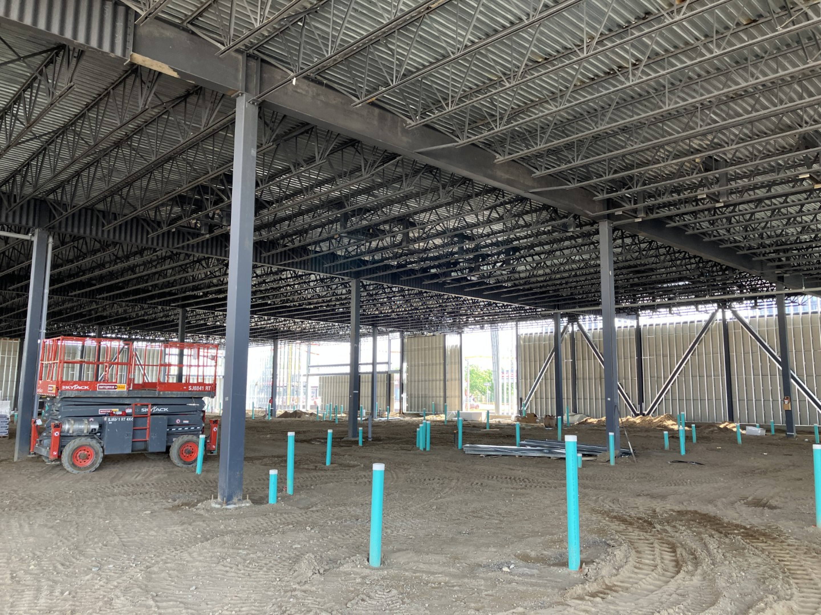 A large unfinished industrial interior with exposed steel roof trusses, steel columns, and compacted dirt flooring. A boom lift sits inside the open space, with blue utility pipe stubs visible across the ground.