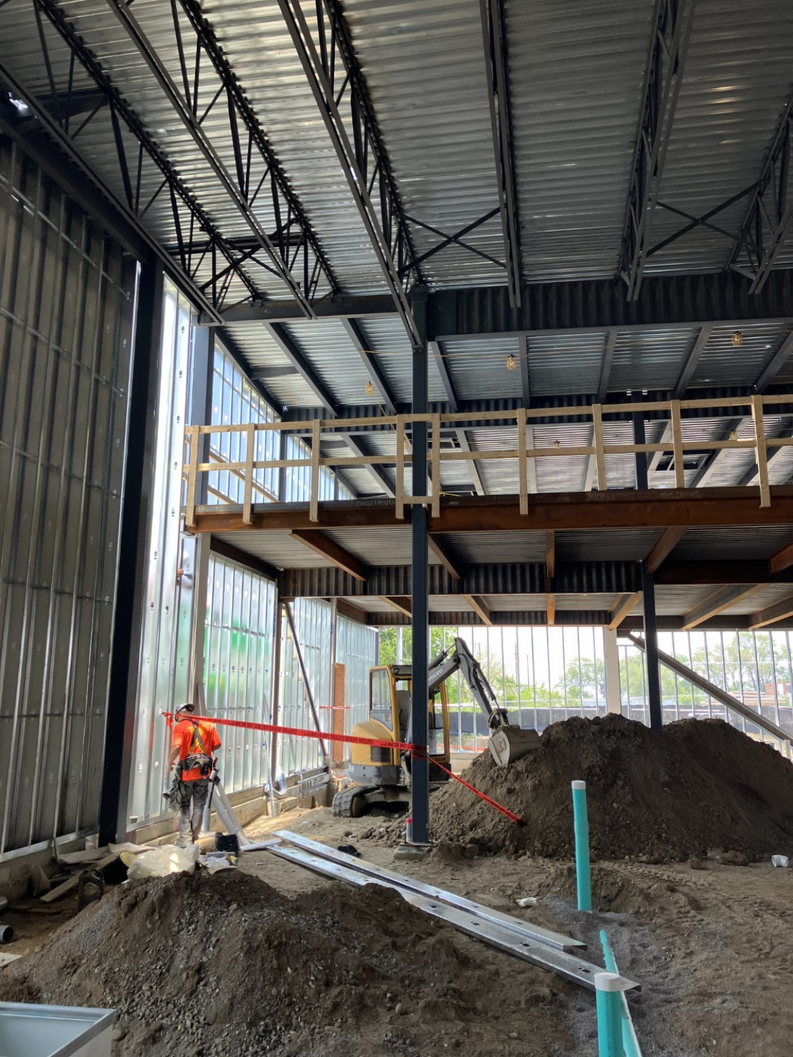 An unfinished industrial interior with exposed steel roof structure and a raised mezzanine platform. Construction materials, soil piles, utility pipes, and a worker operating equipment are visible under natural daylight.