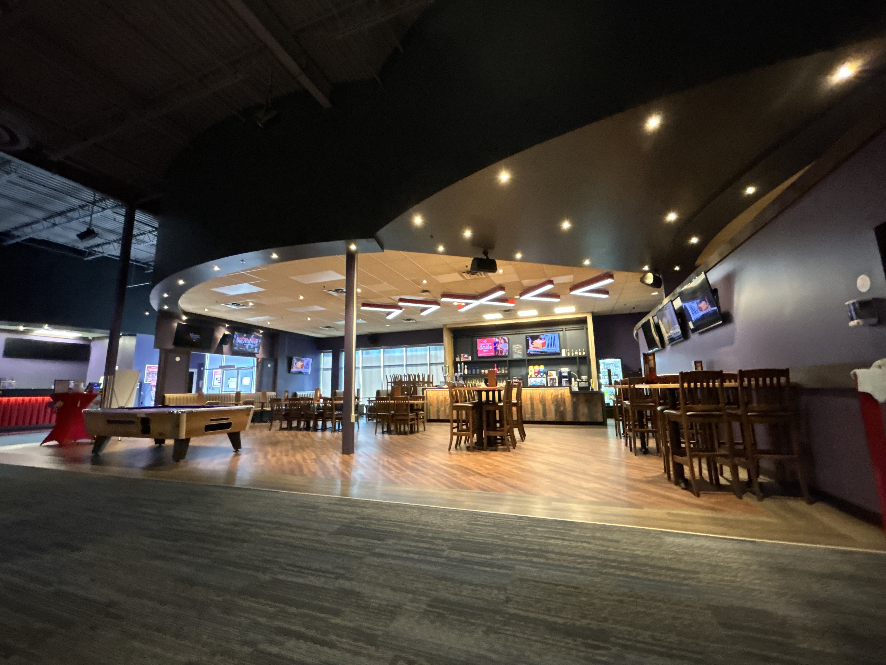 A modern entertainment lounge with a curved ceiling, wood floors, and high‑top tables arranged around a central bar area. Pool tables, wall‑mounted TVs, and built‑in lighting create an open, casual social space.
