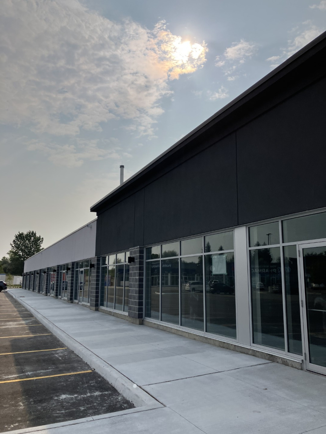 A row of single‑storey commercial storefronts with dark exterior panels and continuous glass windows lines a wide concrete sidewalk. The perspective looks down the building frontage under a partly cloudy sky with sunlight above.
