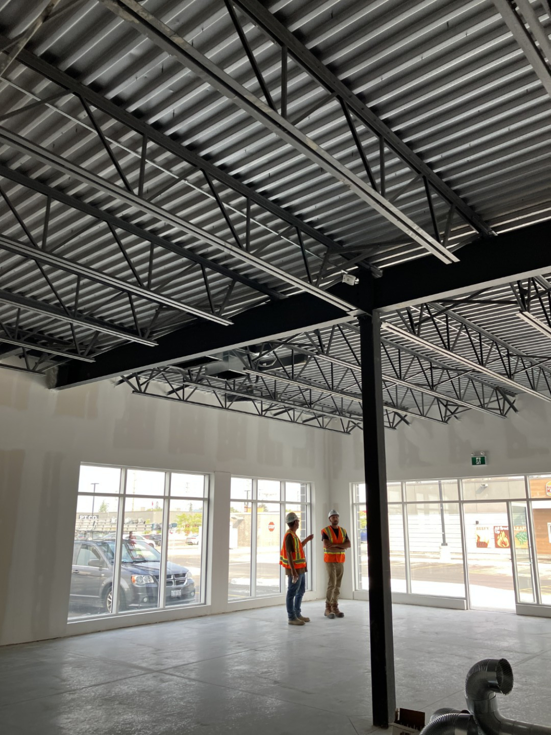 An unfinished commercial interior with exposed metal ceiling decking, steel columns, and concrete floors. Two construction workers stand near large glass storefront doors under bright natural light.