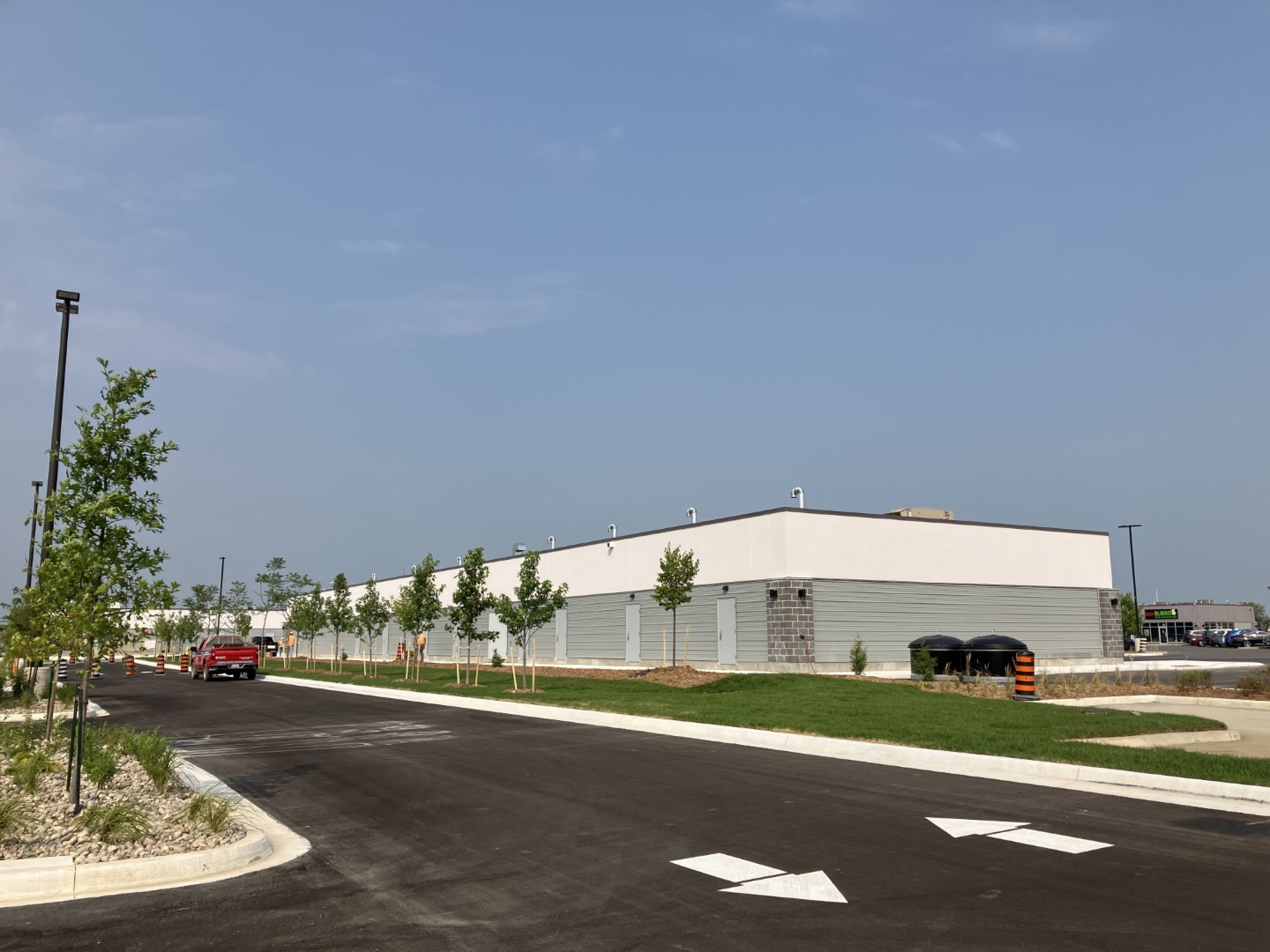 A single‑storey industrial building with white and light gray exterior panels lines a newly paved access road. Young trees, landscaped grass, directional arrows on the pavement, and a clear blue sky frame the scene.