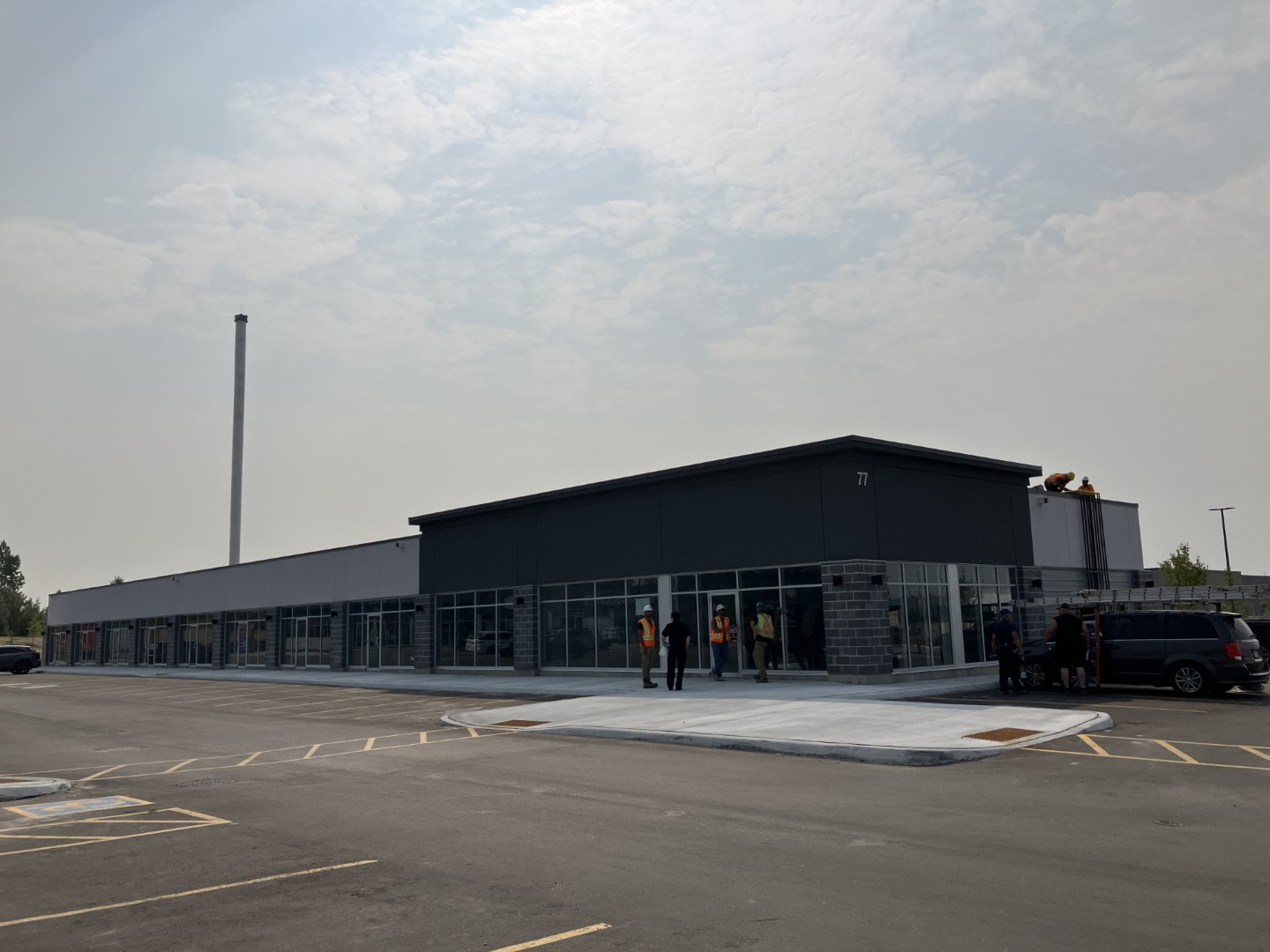 A single‑storey commercial building with a dark gray facade and large glass storefronts faces a wide parking area. A few people stand near the corner entrance beneath a lightly overcast sky.