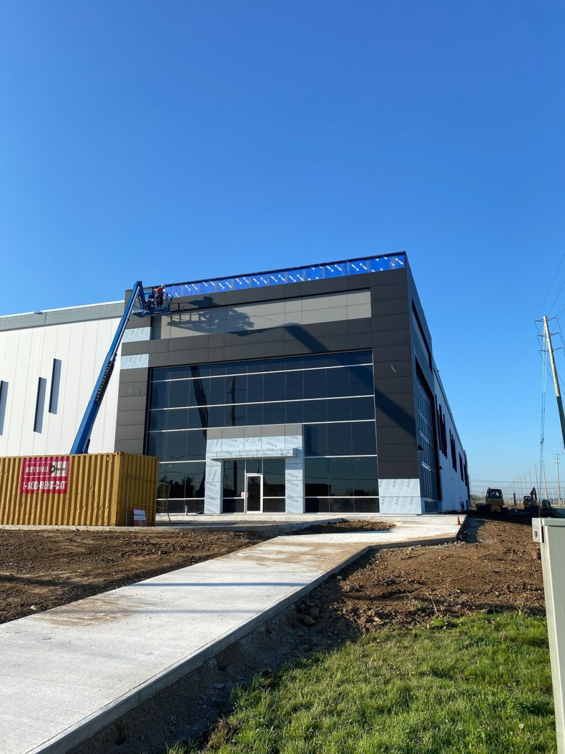 A modern industrial building with a dark gray, glass‑fronted entrance under construction is shown beneath a clear blue sky. A concrete walkway leads to the entrance, with construction equipment and unfinished landscaping visible around the site.