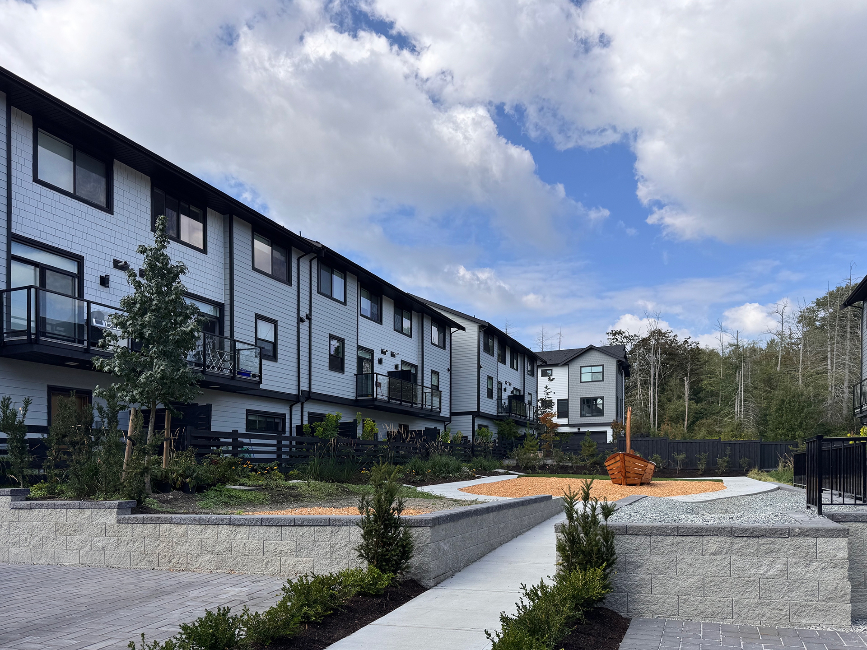 A residential courtyard bordered by modern three‑storey townhomes features paved walkways, landscaped planting beds, and a gravel seating area with wooden benches. Trees and greenery frame the shared outdoor space beneath a partly cloudy sky.