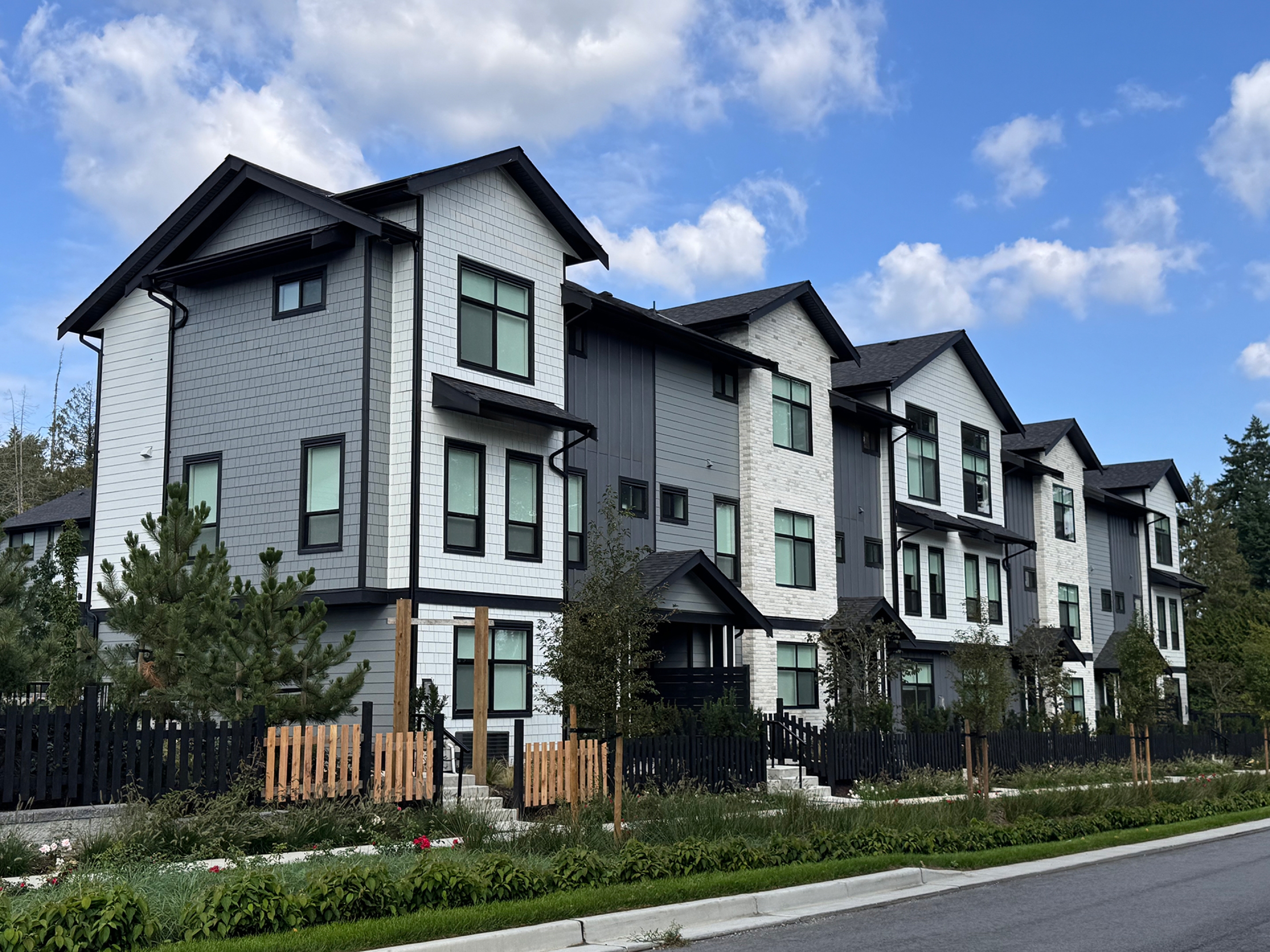 A row of modern three‑storey townhomes with light and dark gray siding lines a landscaped street. Small front yards, young trees, fencing, and a sidewalk sit beneath a bright, partly cloudy sky.