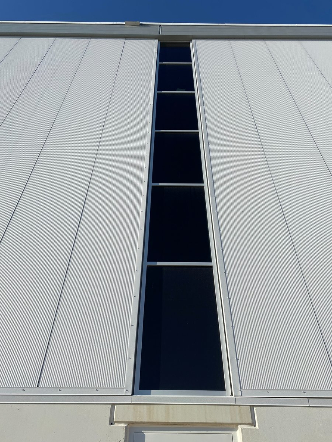 A close-up view of a white metal building facade with vertical ribbed panels and a narrow vertical window strip centered between them. The image is viewed upward, showing clean lines and a clear blue sky above.