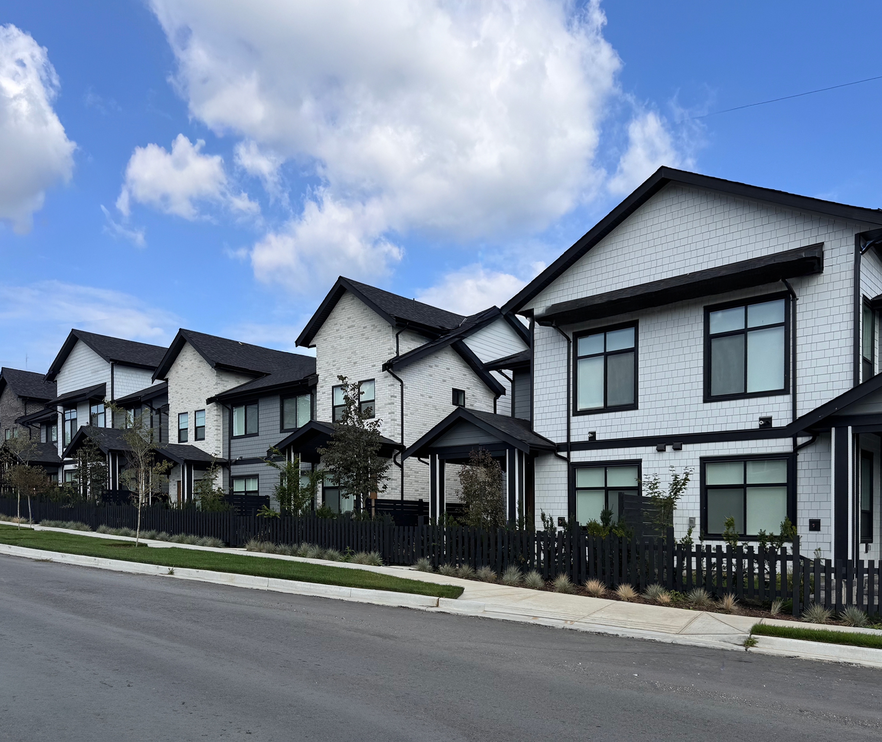 A row of modern three‑storey townhomes with light siding, dark trim, and pitched roofs lines a quiet residential street. Small front yards, black fencing, a sidewalk, and young trees sit beneath a bright, partly cloudy sky.