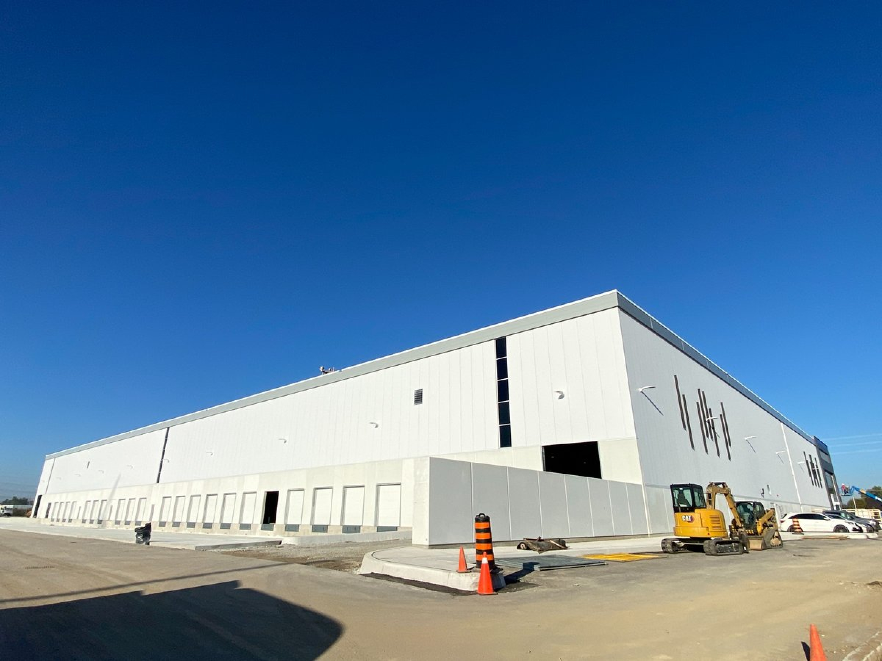 A large industrial warehouse with a white exterior is shown under construction, featuring multiple loading dock doors along one side. Construction equipment, traffic cones, and unfinished pavement are visible under a clear blue sky.