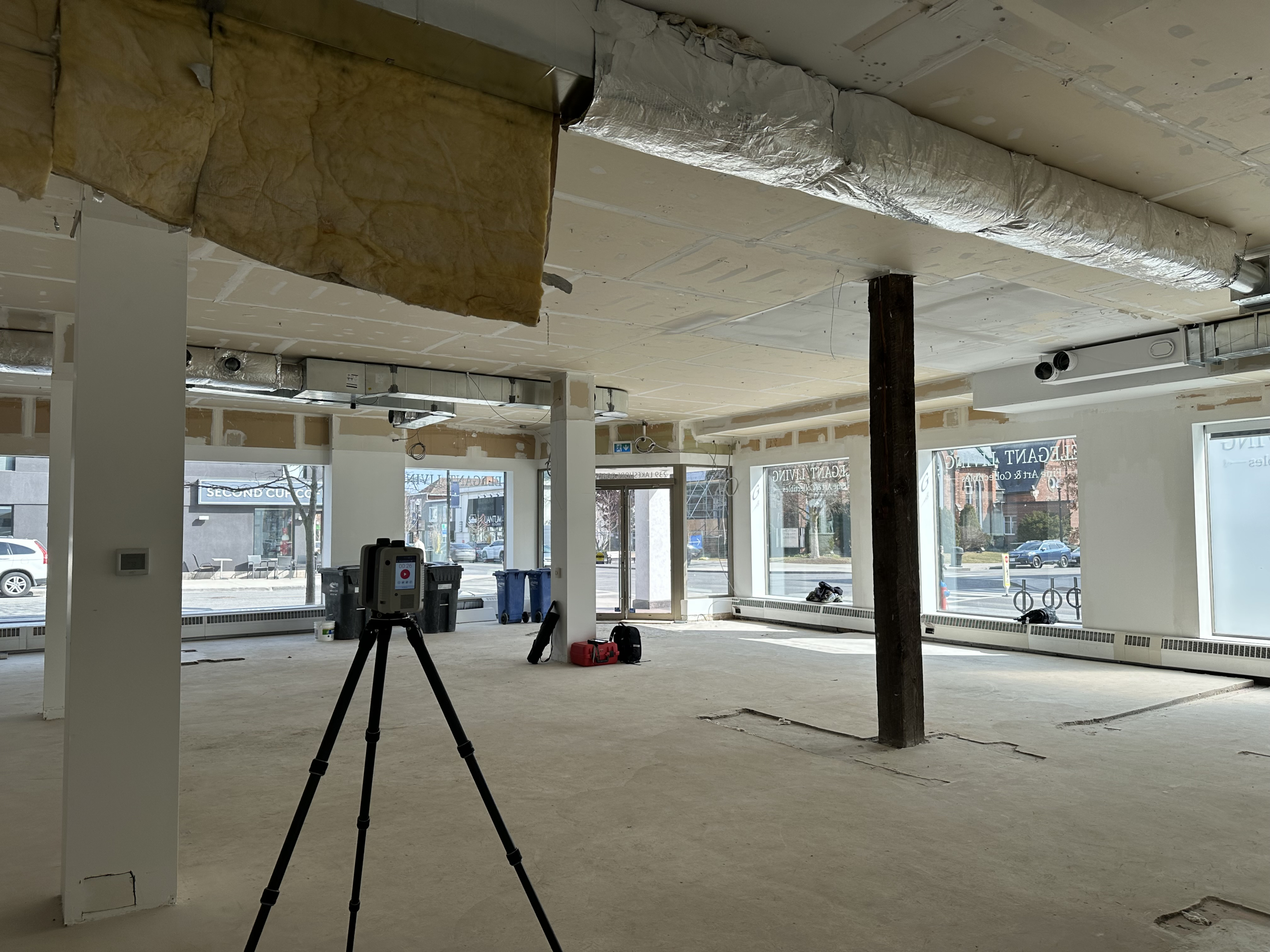 An unfinished commercial interior with exposed ceiling insulation, columns, and concrete floors. A tripod‑mounted 3D scanning device stands in the open space, with large storefront windows along the street-facing walls.
