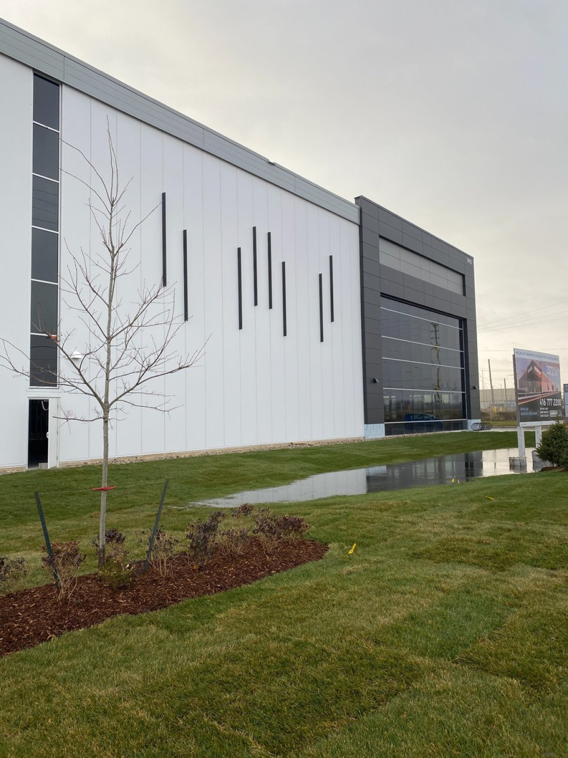 A modern industrial building with a white metal facade and vertical black accent panels sits beside a landscaped grassy area. A dark gray glass‑fronted entrance volume is visible at the far end under an overcast sky.