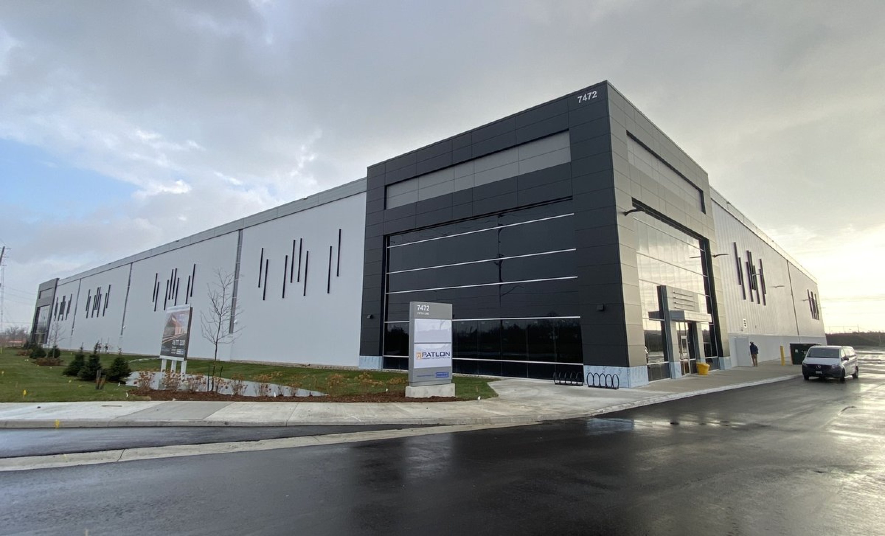 A modern industrial building with a white and dark gray exterior features clean horizontal lines and large glass panels. The structure sits along a paved roadway with landscaped grass, signage near the entrance, and an overcast sky.
