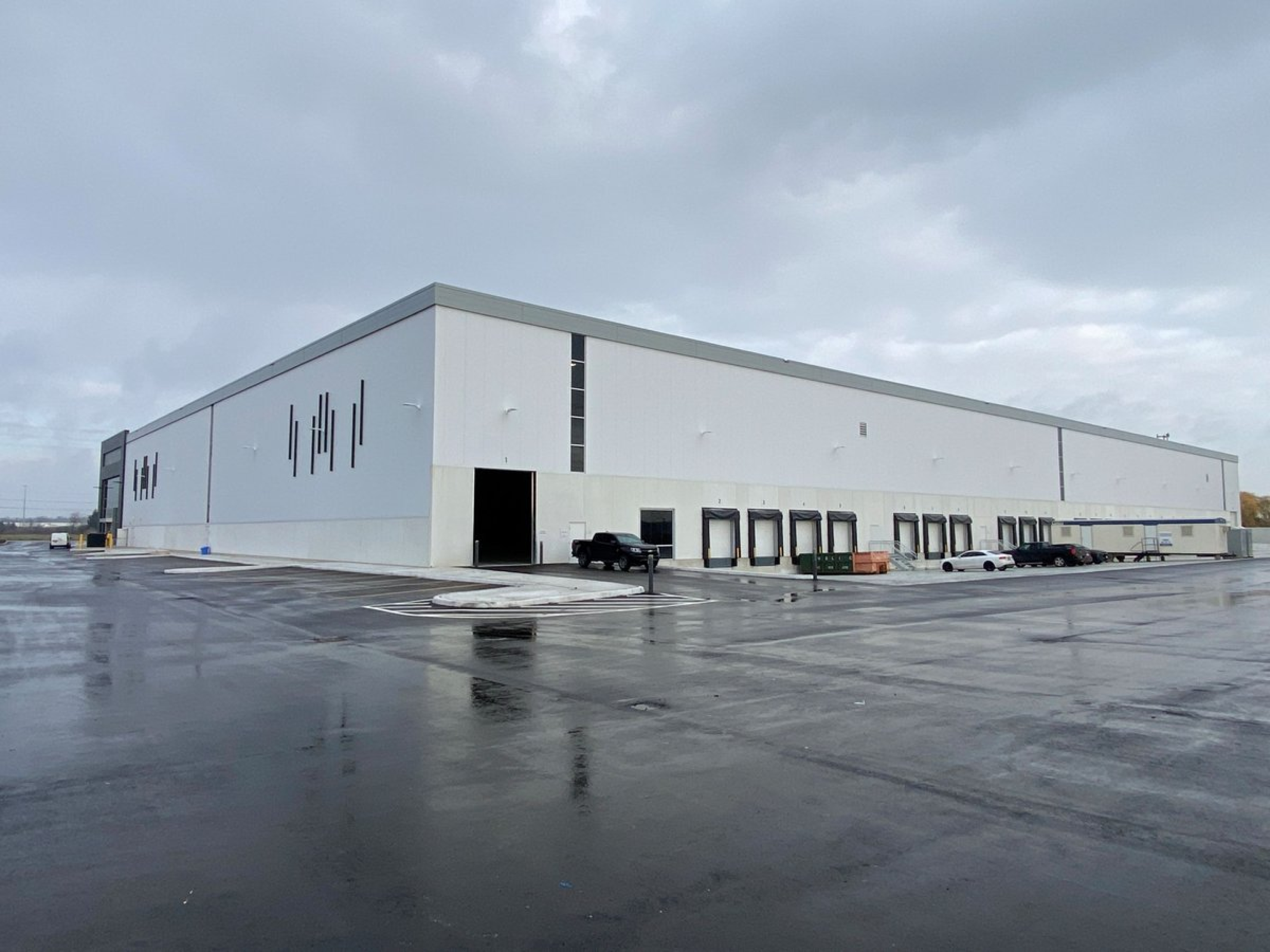 A large white industrial warehouse with multiple loading dock doors lines a wide paved yard. The building features minimal exterior detailing, a few parked vehicles, and wet pavement under an overcast sky.