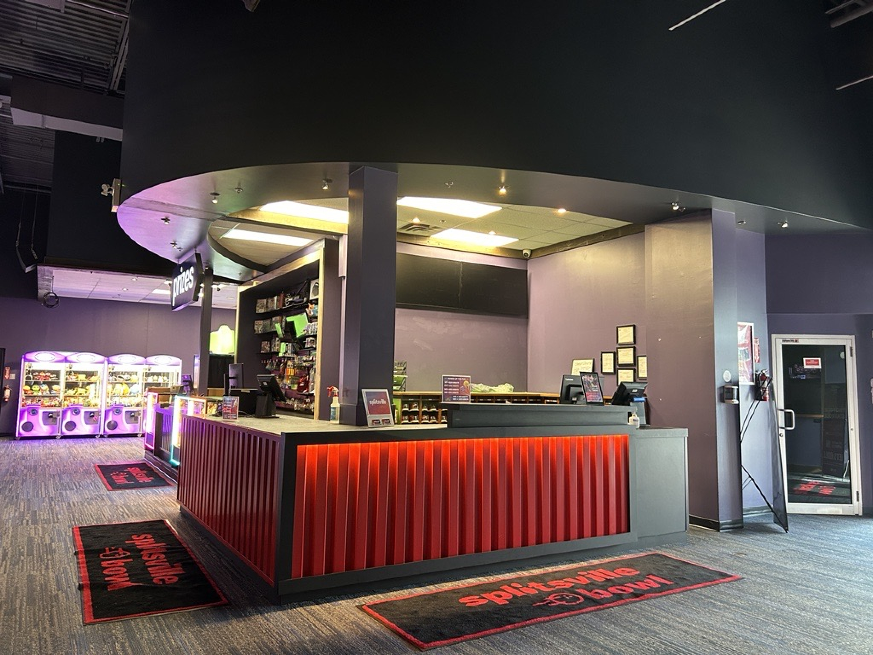 A prize redemption counter inside an entertainment venue with a curved overhead canopy and a red‑paneled front desk. Digital monitors, shelving, and colorful arcade machines are visible in the background under bright lighting.