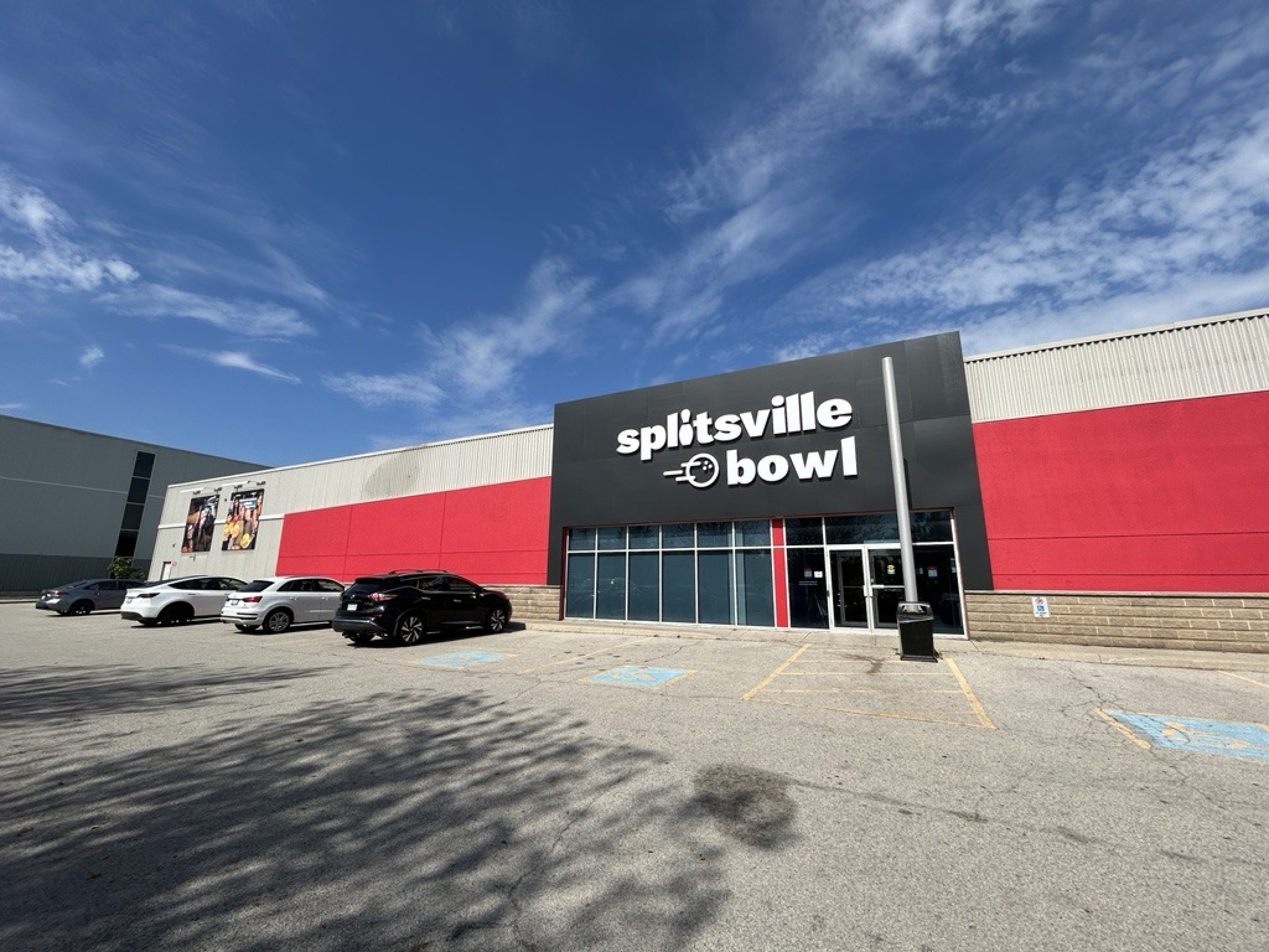 A Splitsville Bowl exterior with a wide red facade and a black sign above the main entrance. Large windows face a parking lot with several cars, under a bright blue sky with wispy clouds.