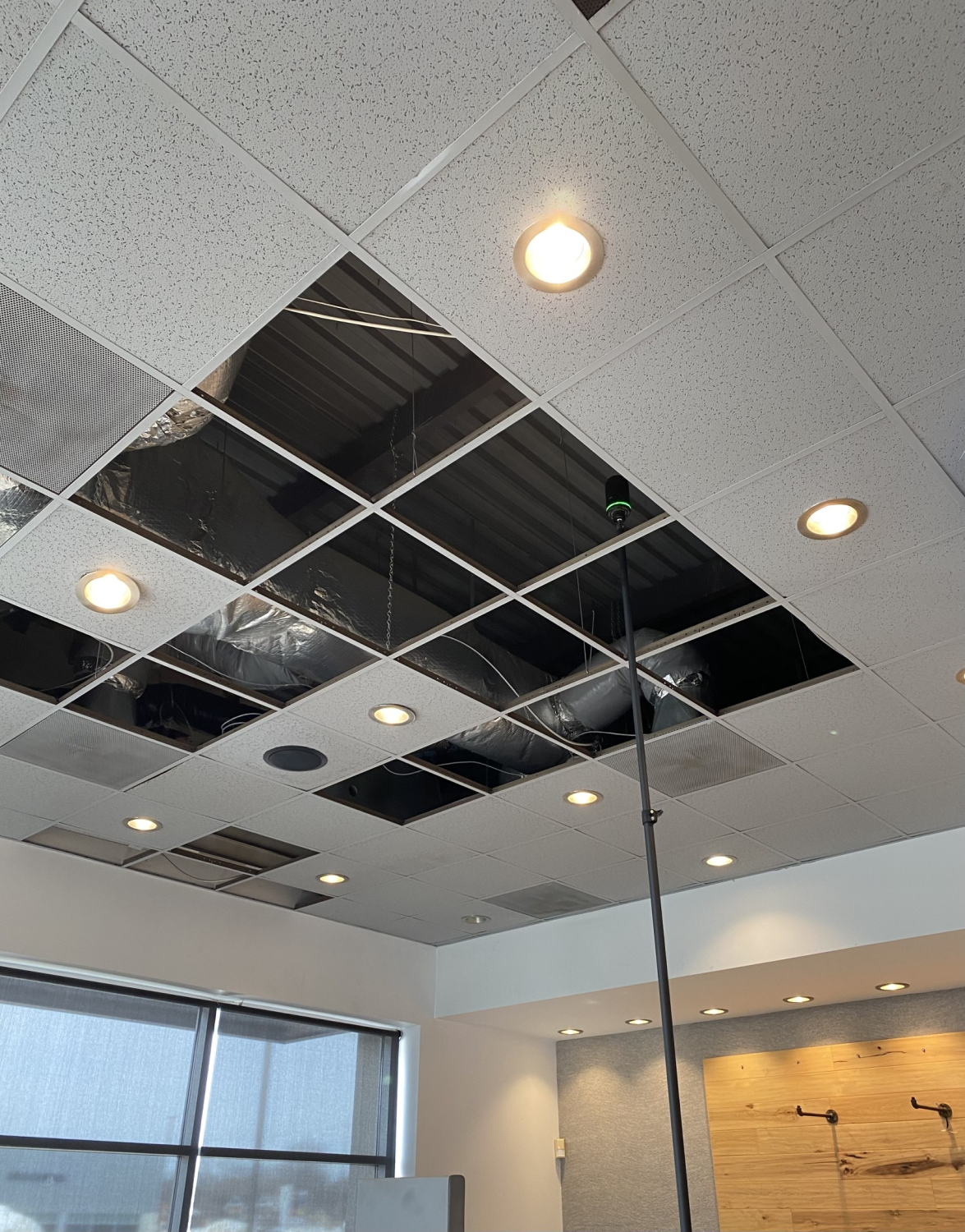 A restaurant interior ceiling with a partially open drop‑ceiling grid showing exposed ductwork and insulation above. Recessed lights are set within the ceiling tiles, with windows and wall finishes visible below.