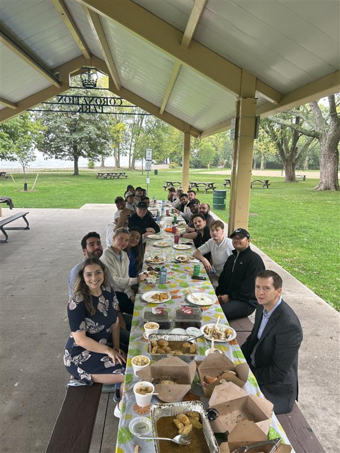 A long table of coworkers sits under a covered park pavilion, sharing a group meal at an outdoor picnic. Green grass, trees, and picnic tables surround the gathering in the open park setting.