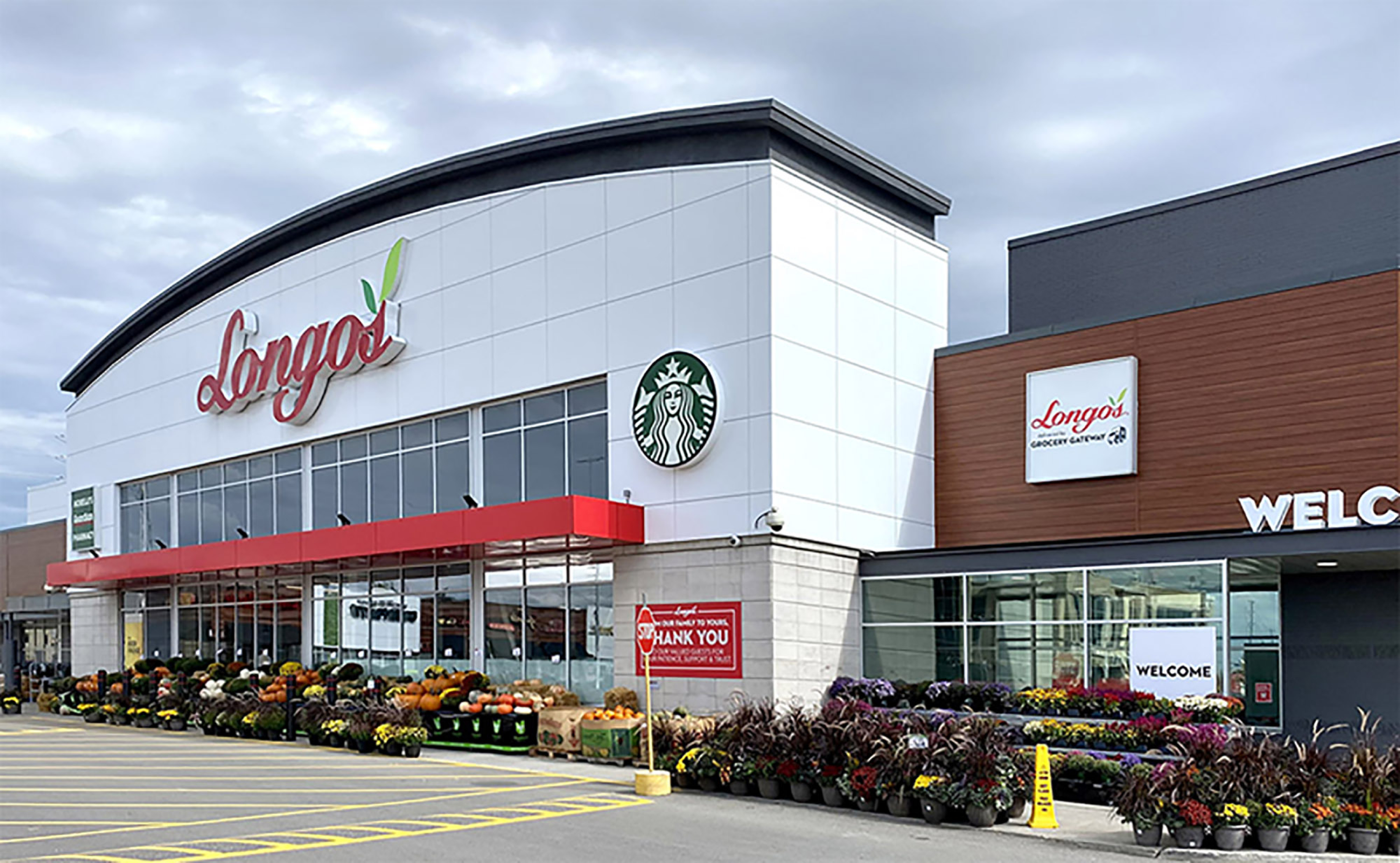 A Longo’s grocery store exterior with a curved white facade and red “Longo’s” sign above large front windows. A Starbucks logo is mounted on the building, with outdoor plant displays and parking spaces in front.