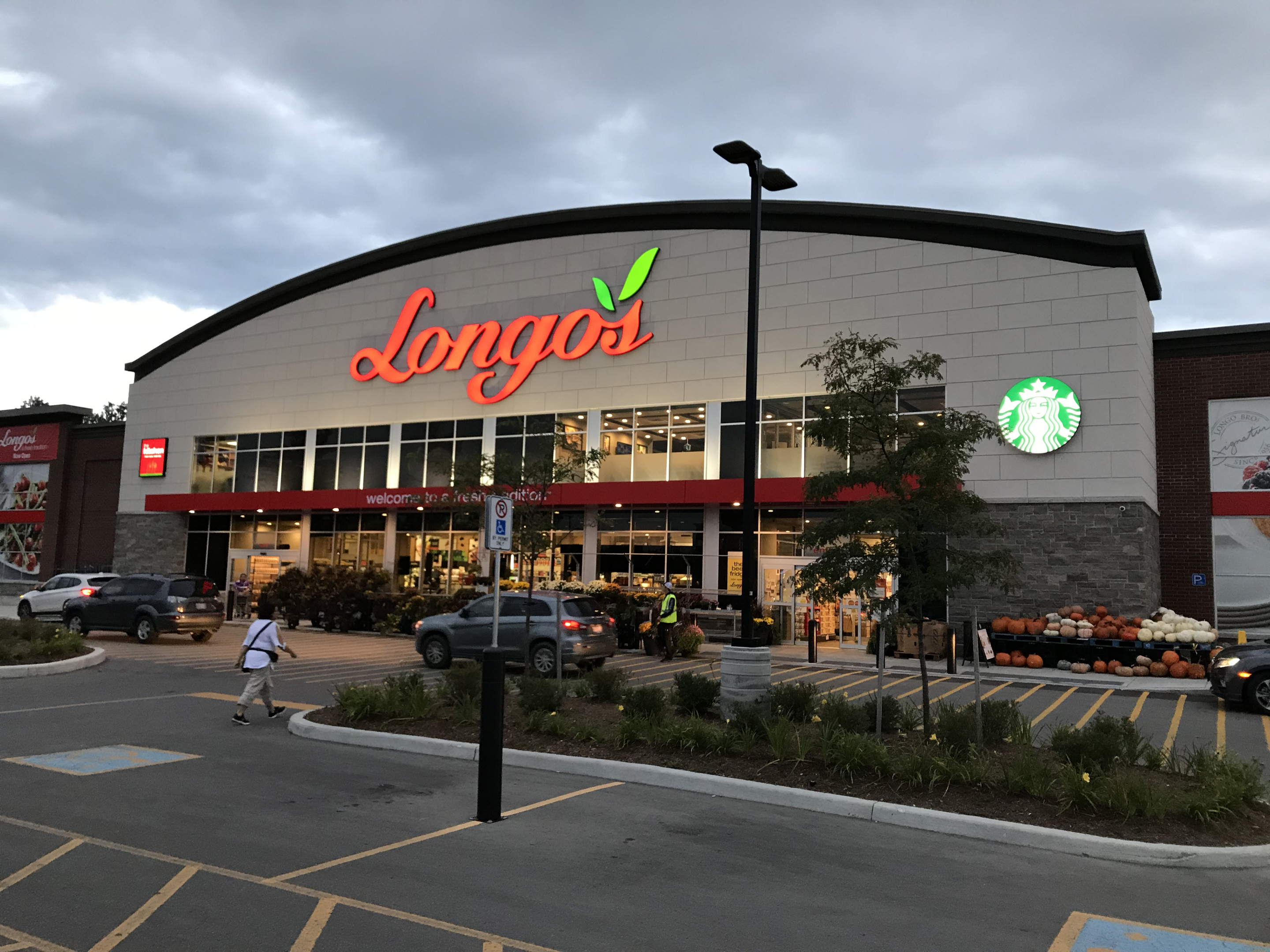 A Longo’s grocery store exterior with a curved white facade and red “Longo’s” sign above large front windows. A Starbucks logo is visible on the building, with cars, pedestrians, and outdoor produce displays near the entrance.