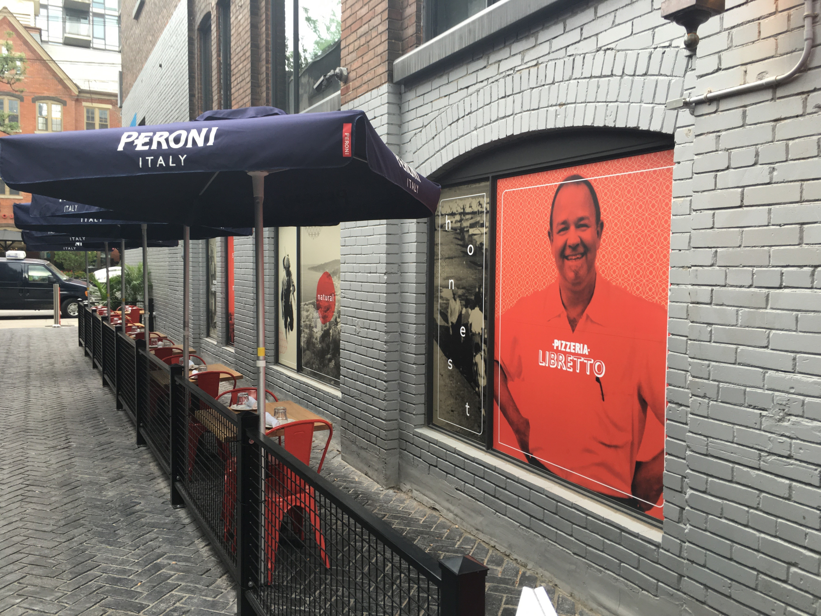 An outdoor restaurant patio set along a brick-lined alley, with tables and red chairs enclosed by a black metal fence. A dark blue umbrella labeled “PERONI ITALY” shades the seating beside a wall featuring large restaurant posters.