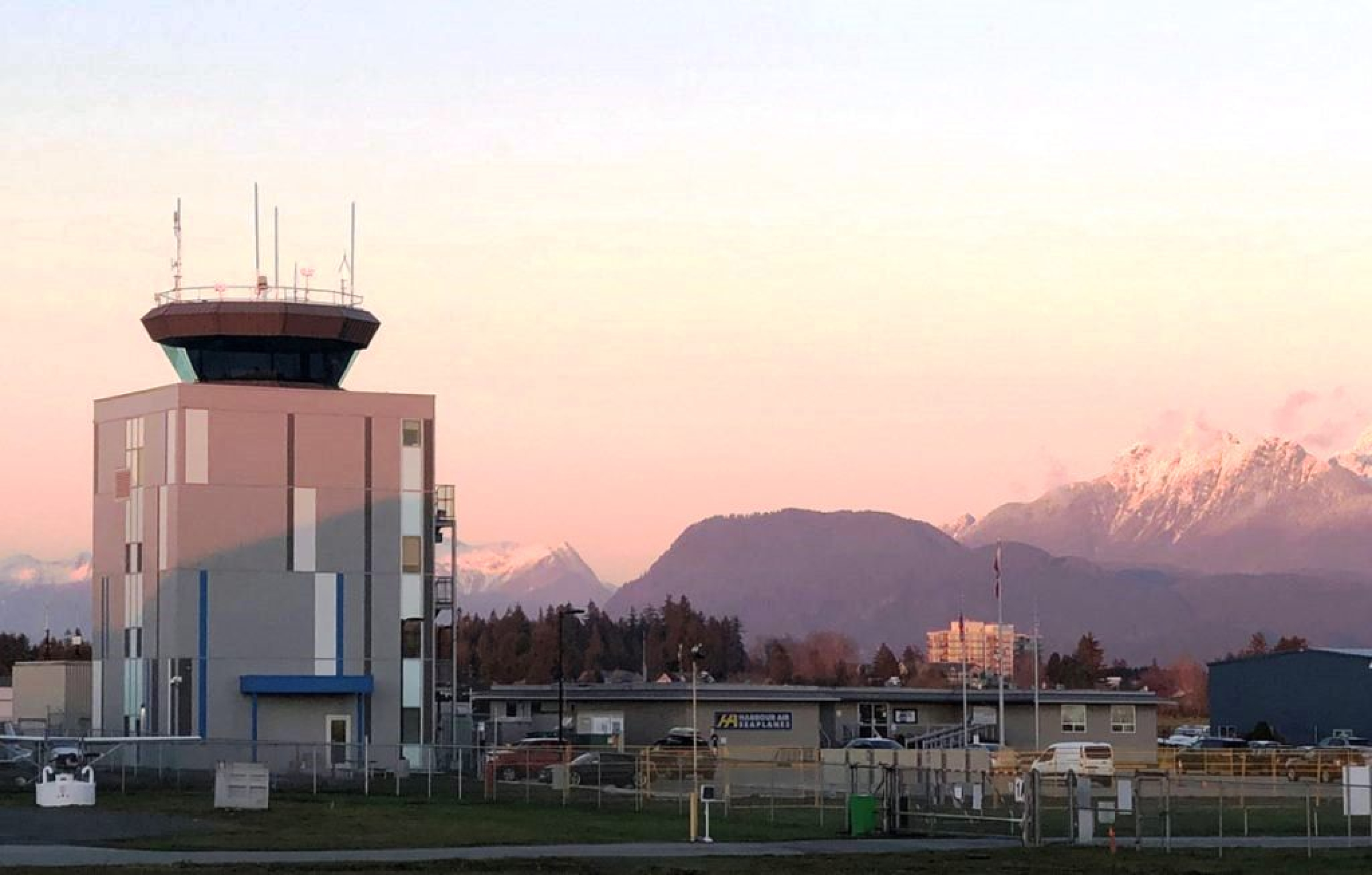 A NAV CANADA air traffic control tower with a gray and blue exterior stands within a fenced operations site. Forested hills and snow‑capped mountains rise in the background under a soft sunset sky.