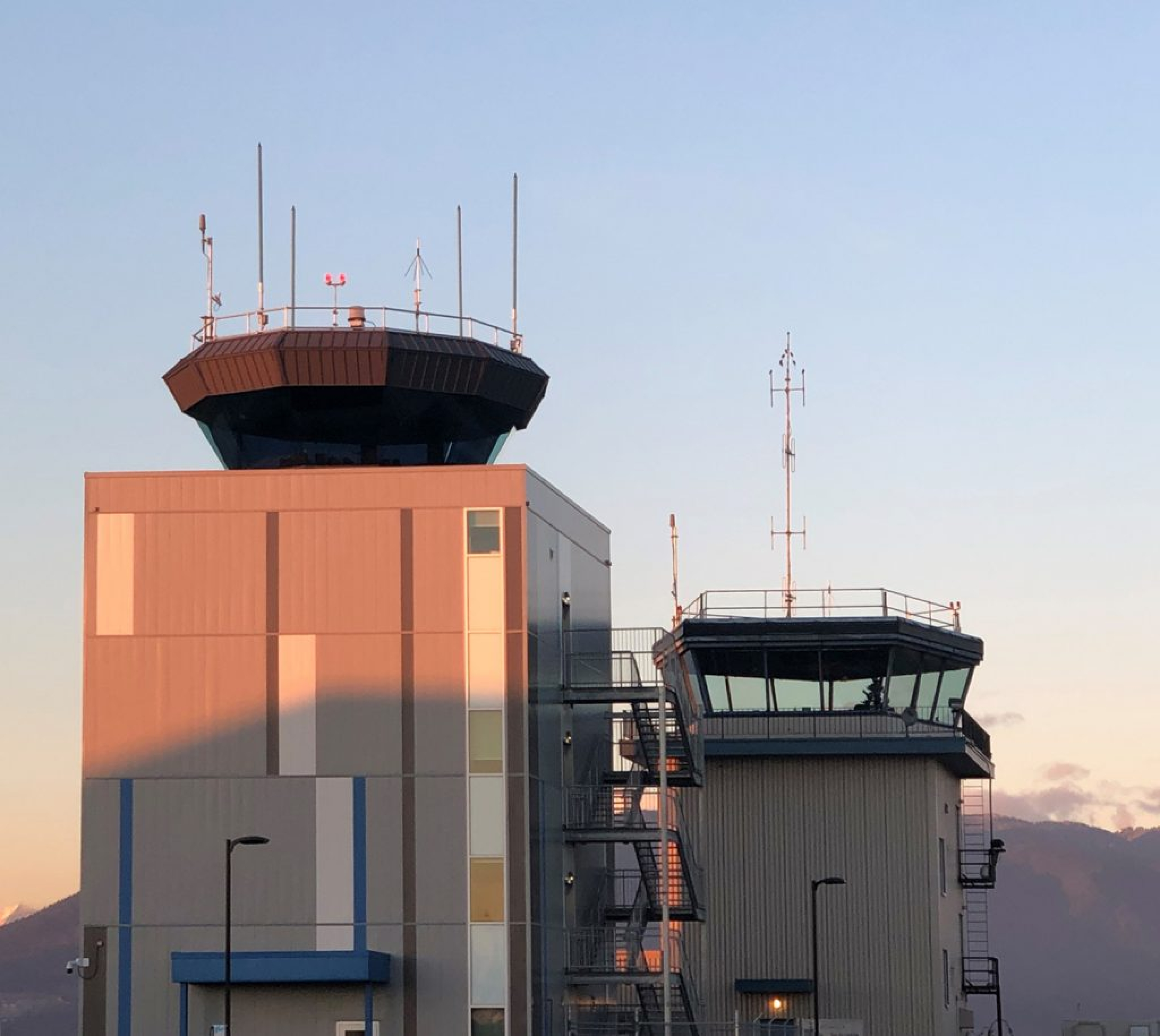 Two NAV CANADA air traffic control towers with antenna‑topped control rooms stand side by side at sunset. Warm light highlights the gray and blue metal structures against a clear sky and distant mountains.