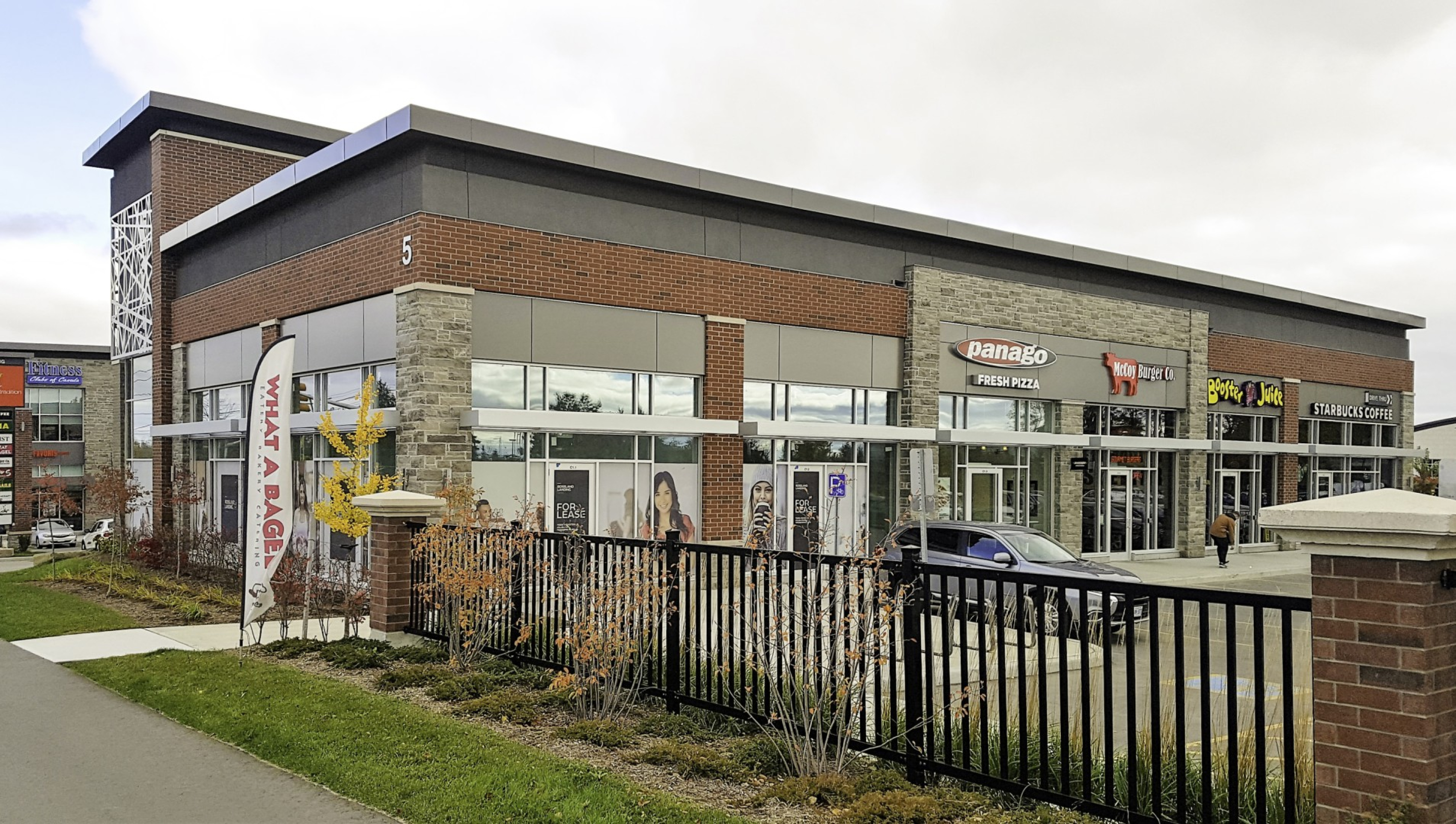 A modern two‑storey retail building with brick, stone, and metal exterior finishes, housing multiple storefronts with large glass windows. A parking area, sidewalk, and fencing sit in the foreground beneath an overcast sky.