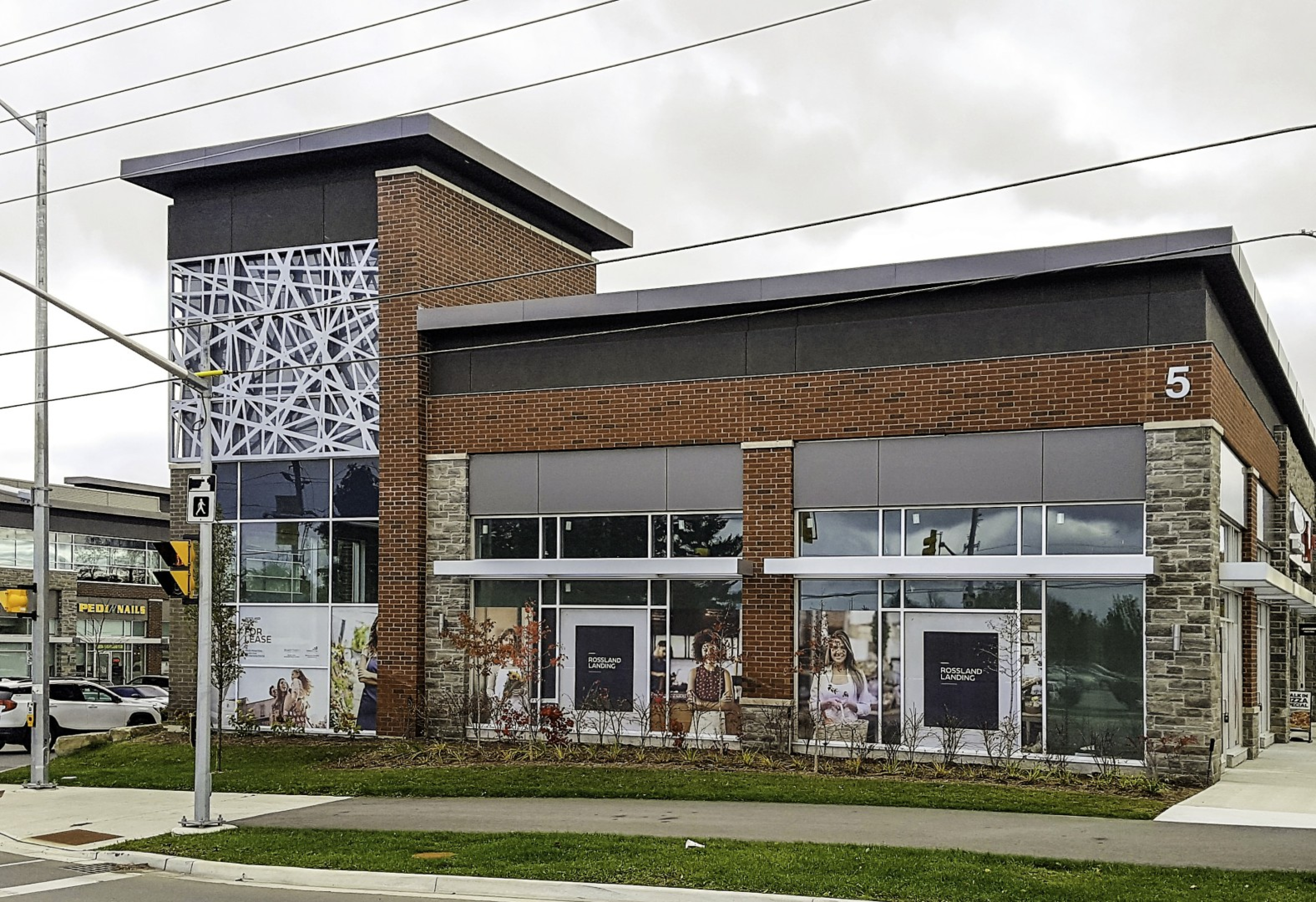 A corner view of a modern two‑storey retail building with brick and stone walls, glass storefronts, and a decorative metal screen on a tower feature. Sidewalks, landscaping, and a street intersection appear in the foreground under an overcast sky.