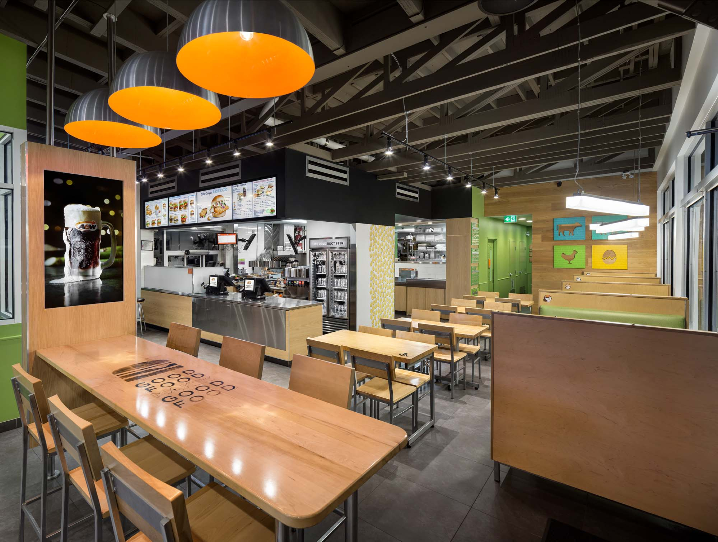 Modern fast-food restaurant interior with light wood tables, booth seating, and orange pendant lights beneath an open ceiling. The ordering counter and illuminated menu boards are visible in the background.