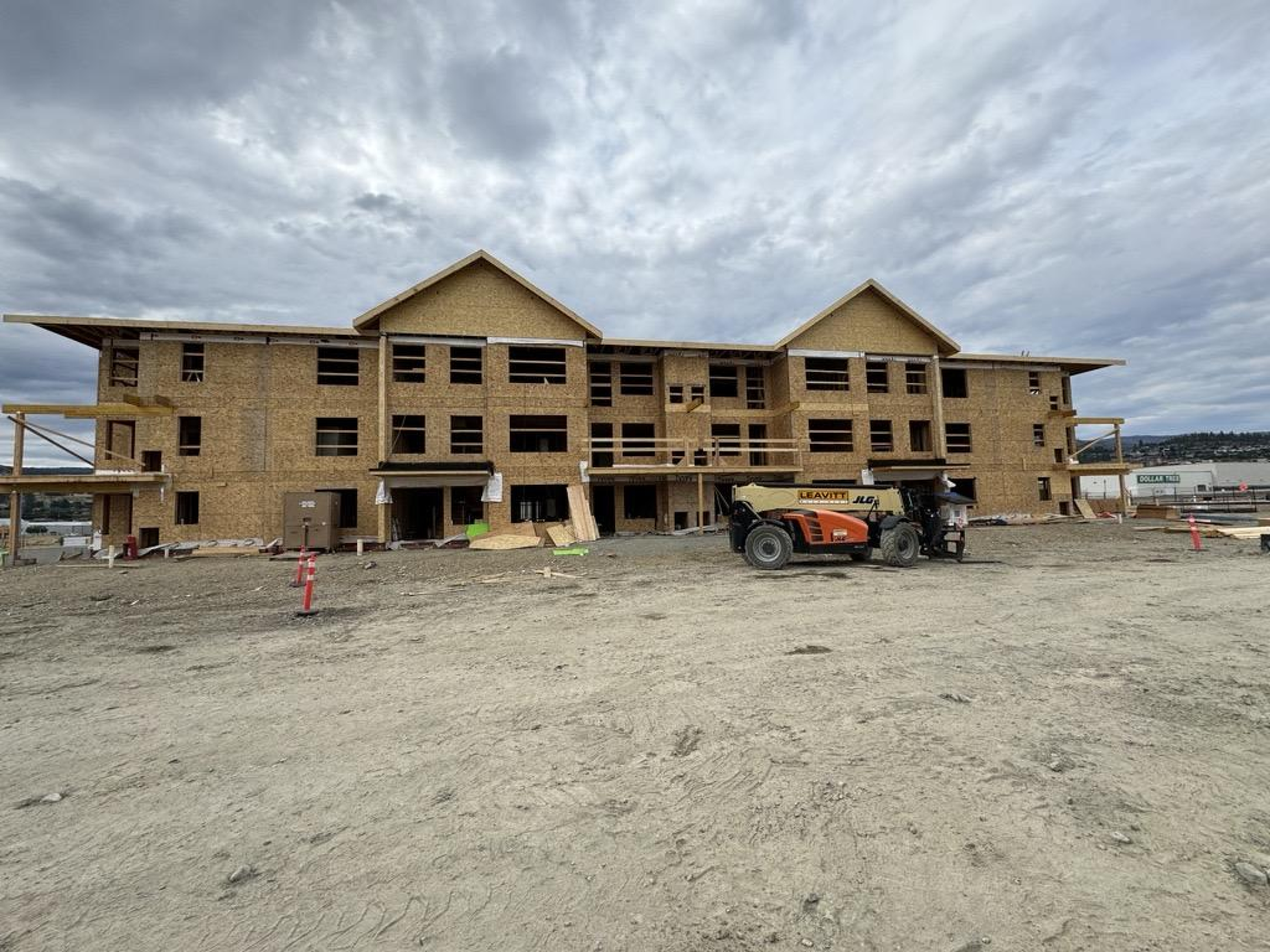 A three‑storey residential building under construction with exposed wood framing and unfinished window openings. Construction equipment and materials sit on a dirt site in front of the structure beneath an overcast sky.