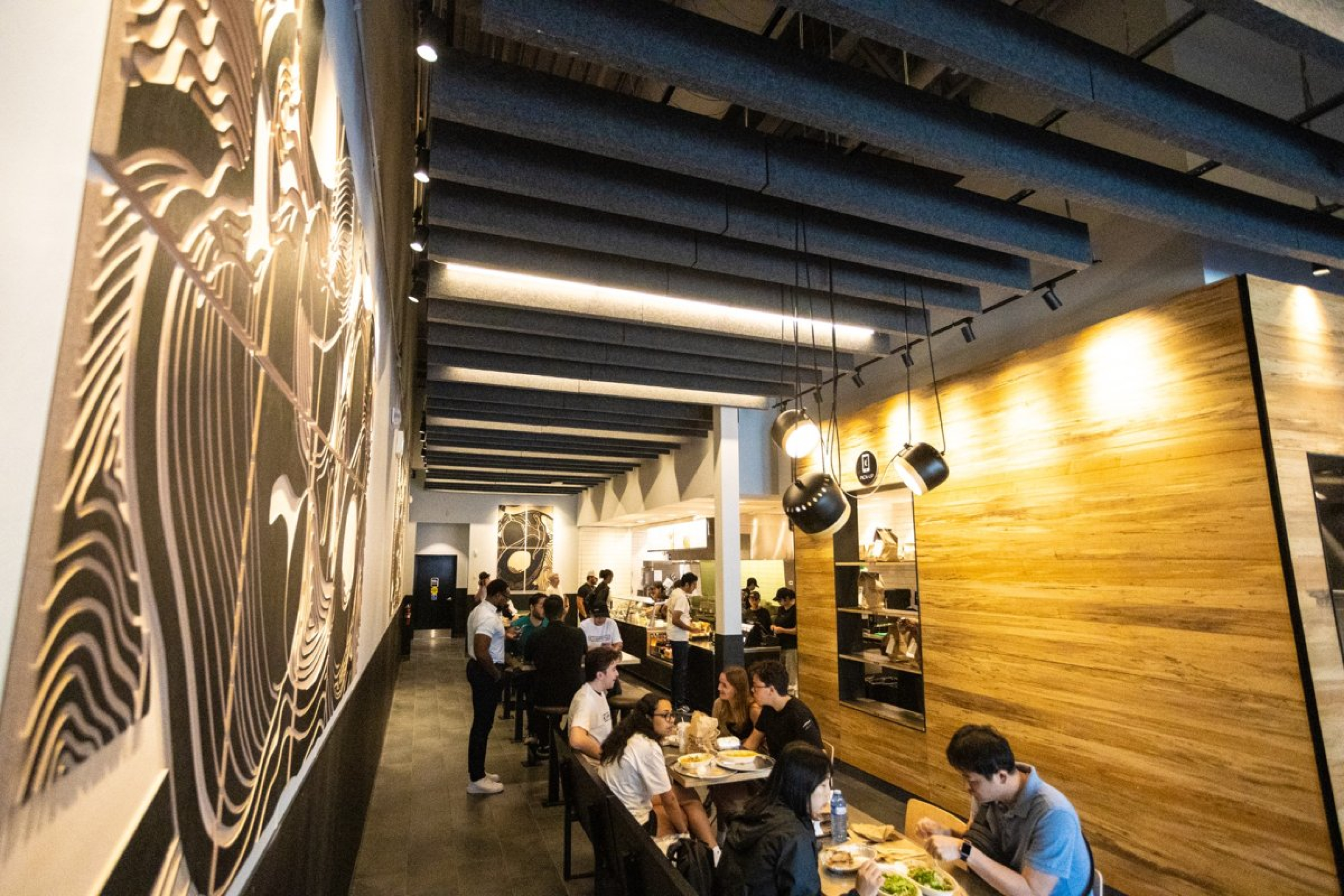 A busy restaurant interior with long tables and bench seating, where patrons are dining along a narrow aisle. Wood-paneled walls, exposed ceiling beams, and hanging lights create a warm, modern atmosphere.