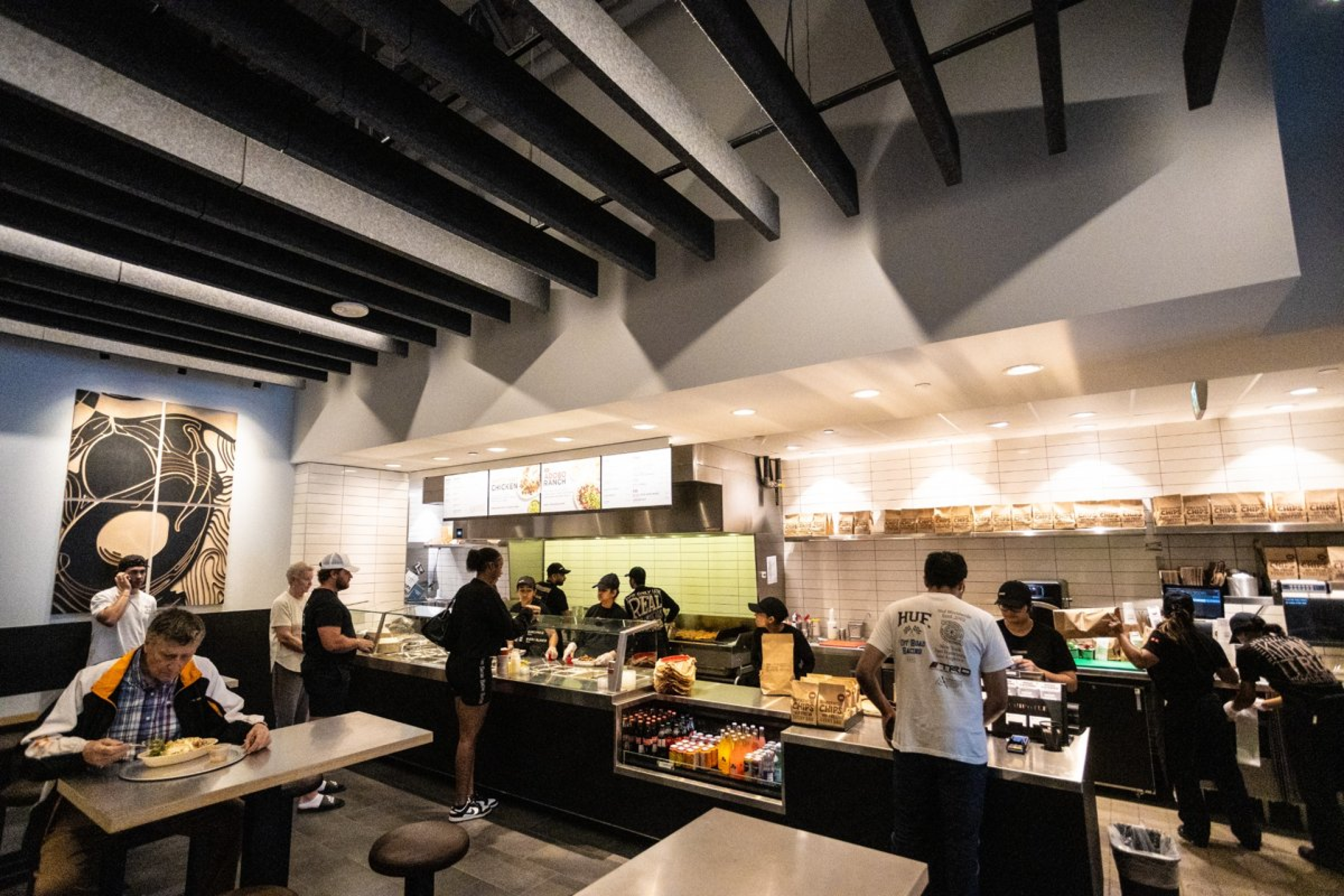 A restaurant interior with customers seated at tables and others ordering at a long service counter. Exposed ceiling beams, menu boards, glass display cases, and bright lighting create a modern, busy dining environment.