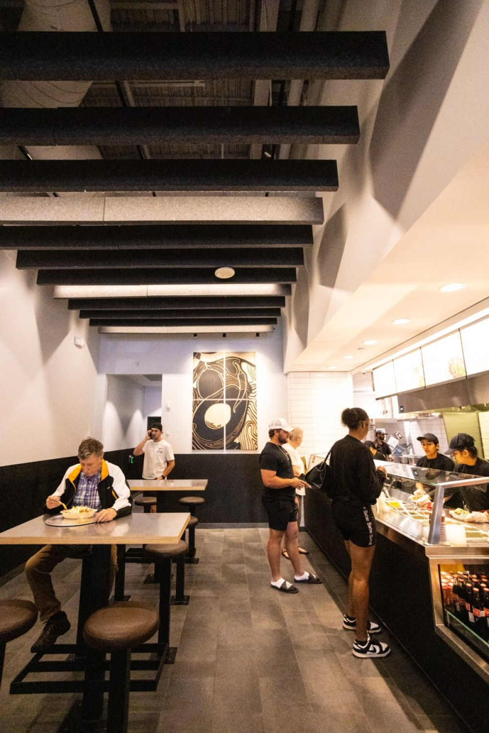 A restaurant interior with customers seated at tables and others ordering at a service counter. Exposed black ceiling beams, neutral walls, glass display cases, and overhead lighting create a modern, casual setting.