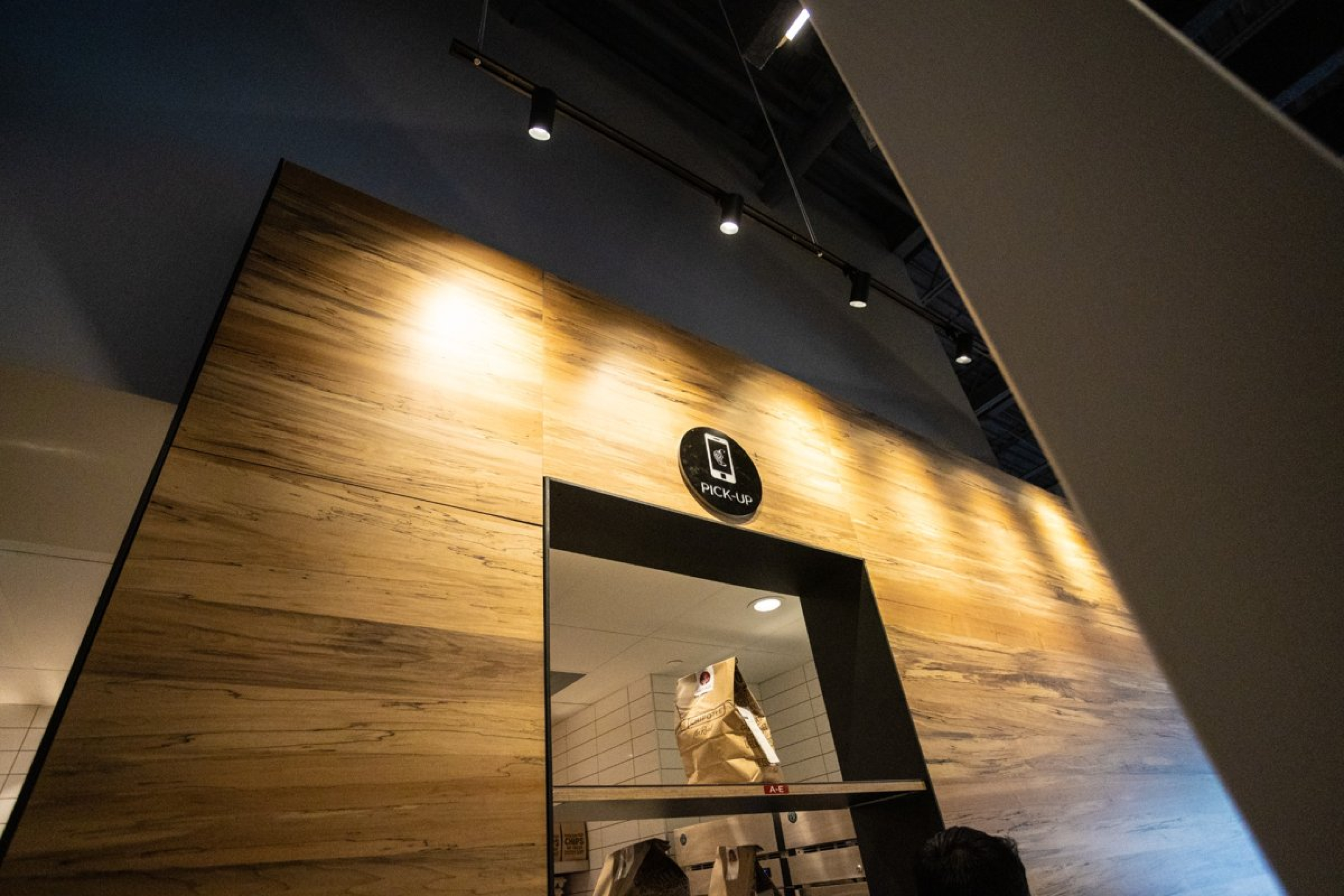 A restaurant pickup window set into a wood-paneled wall, with a paper takeout bag visible inside the opening. Overhead track lights illuminate the area, and a small “Pick Up” sign is mounted above the window.