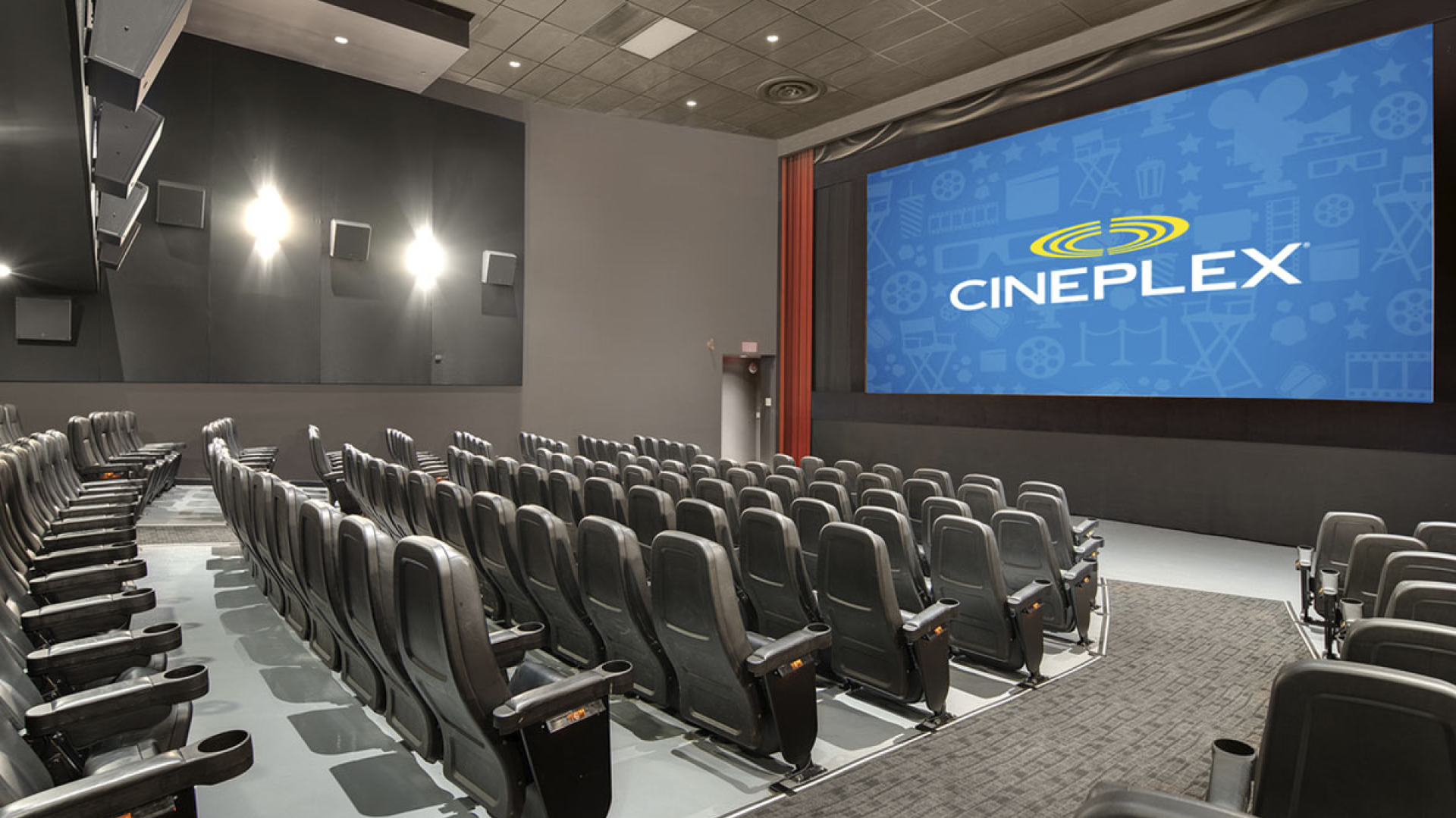 A Cineplex movie theatre auditorium with rows of black reclining seats facing a large screen displaying the Cineplex logo. Wall lights, tiered seating, and wide aisles create a spacious, modern cinema setting.