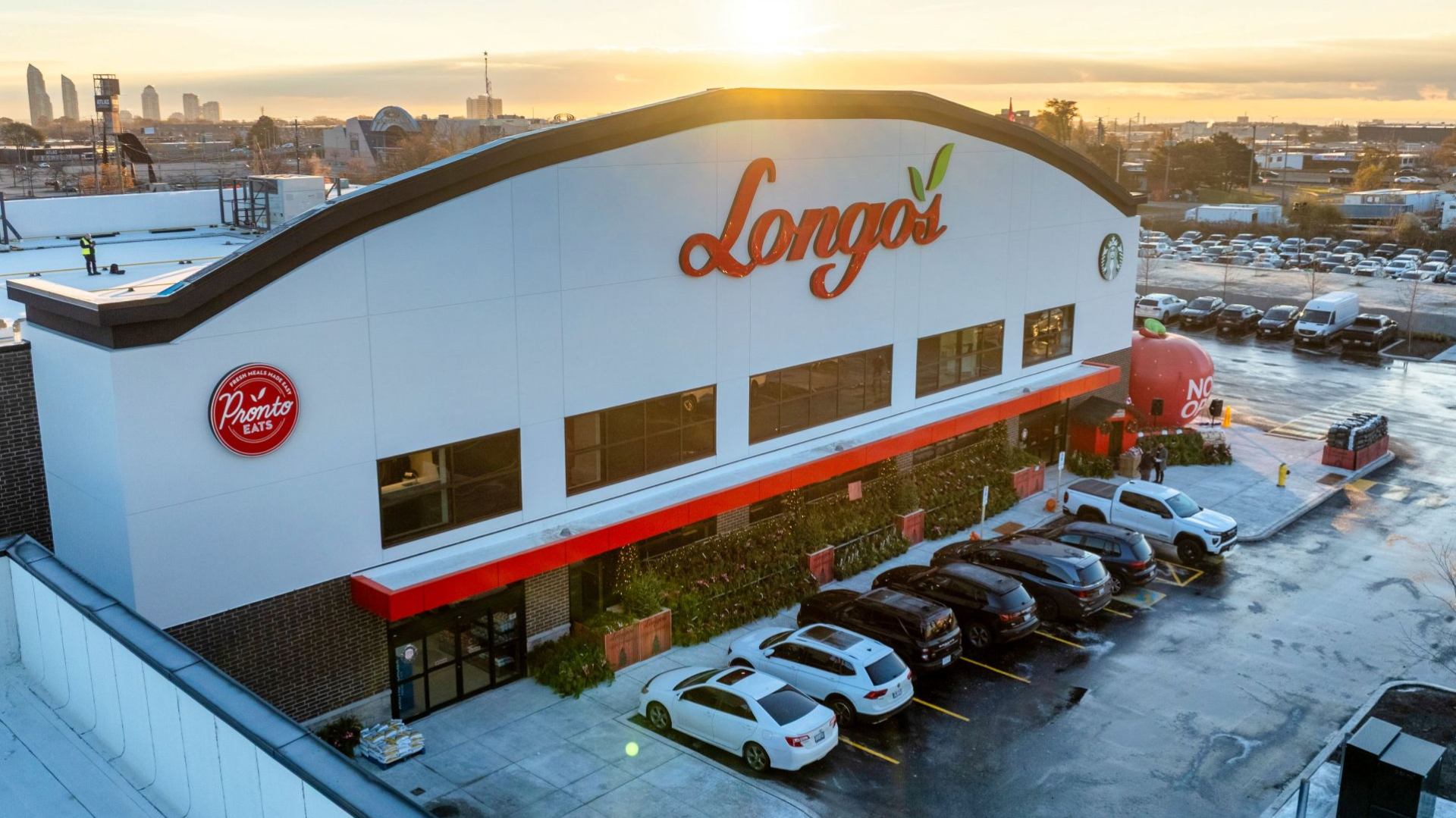 An aerial view of a Longo’s grocery store with a curved white facade and red signage, surrounded by a parking lot with parked cars. Snow and winter conditions are visible, with the city skyline in the background at sunset.